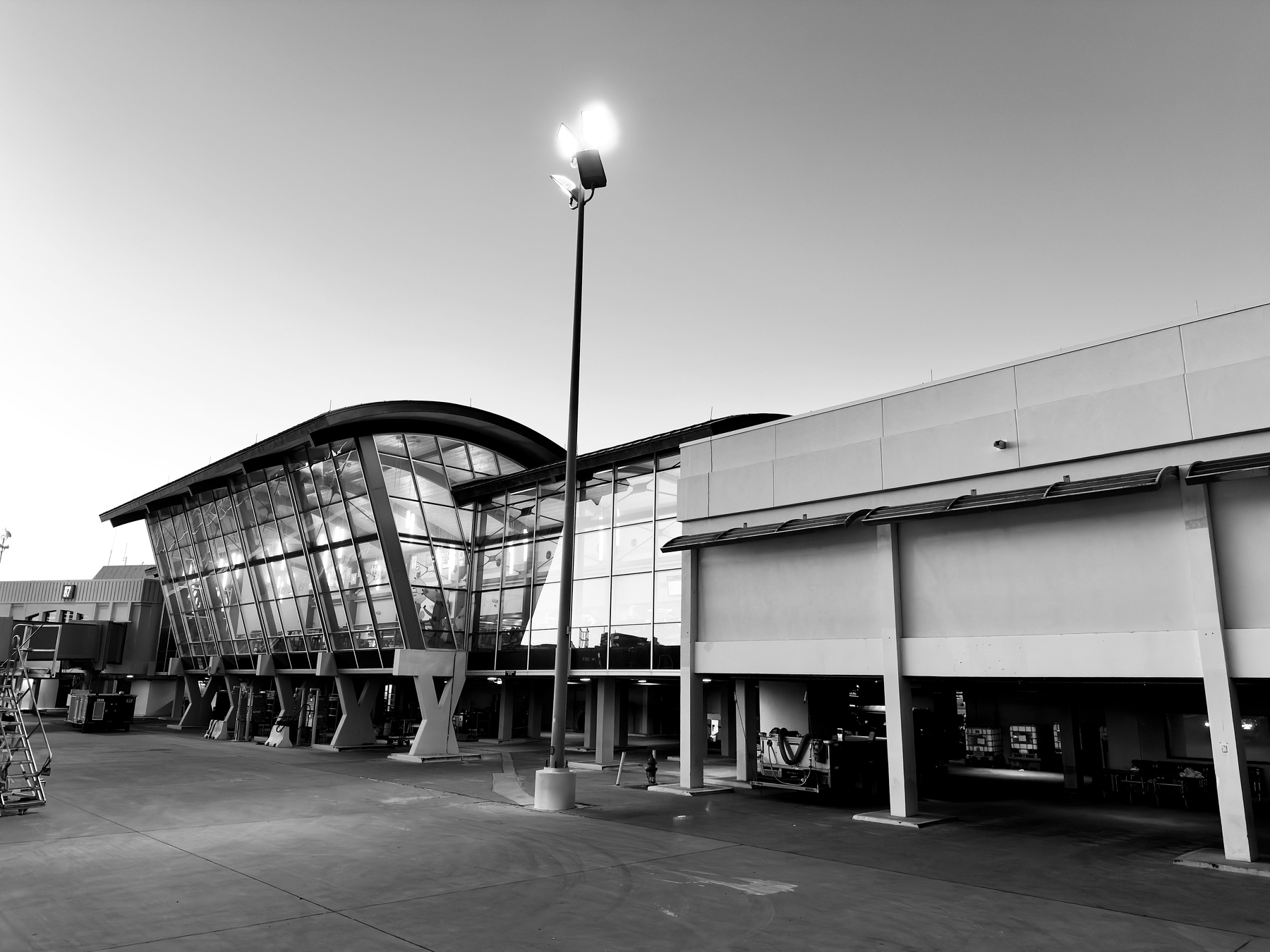 Auto-generated description: A modern airport terminal with large glass windows and an adjacent parking or service area is illuminated by tall light poles during dusk or dawn.