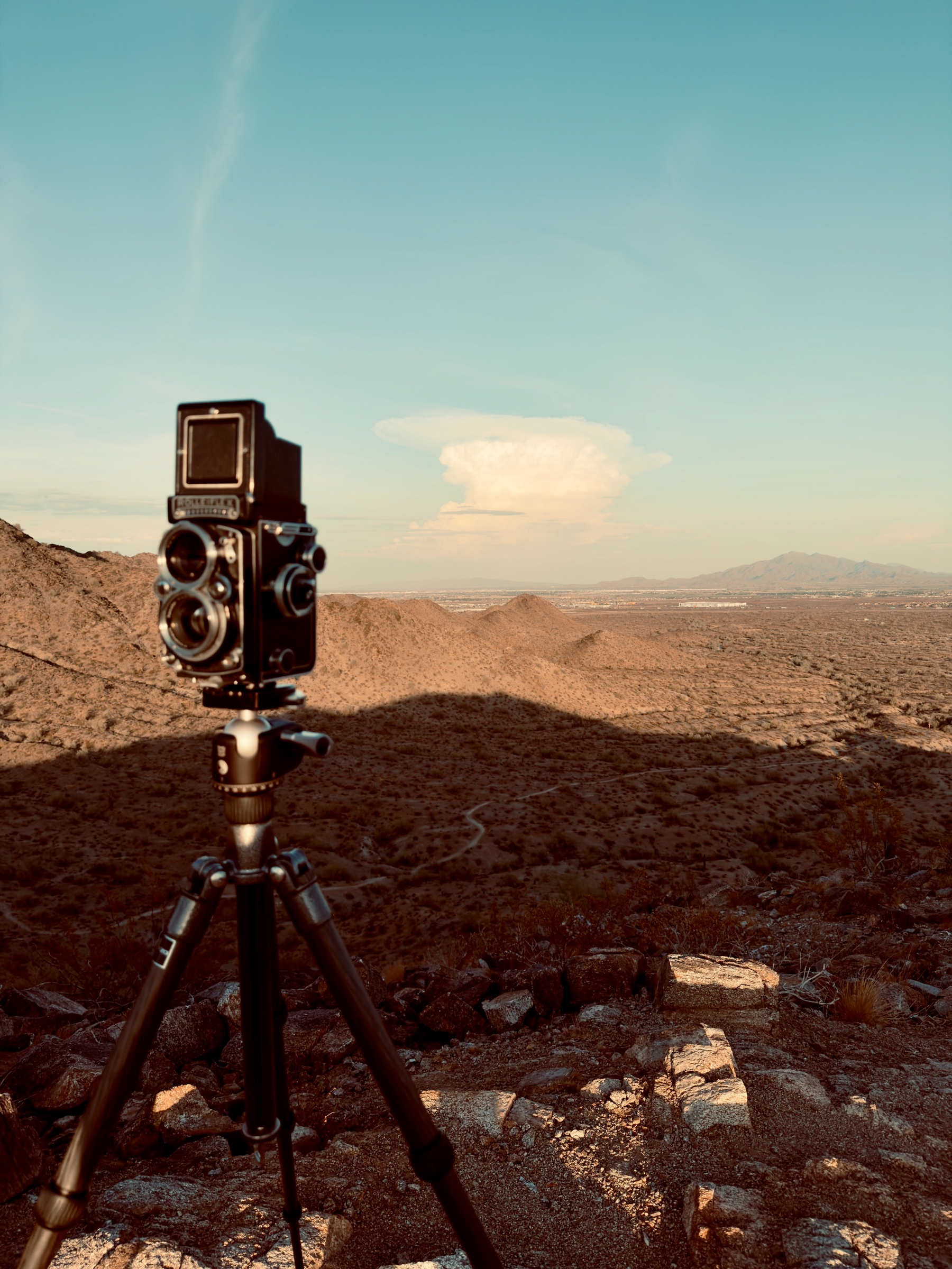 Auto-generated description: A vintage twin-lens reflex camera is mounted on a tripod capturing a wide desert landscape under a clear sky with a distant cloud formation.