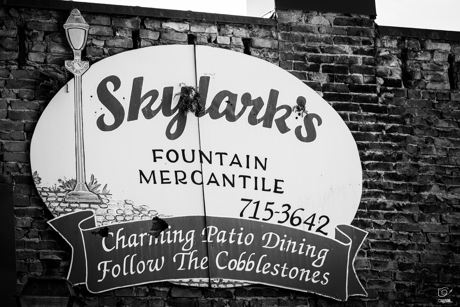 Auto-generated description: A vintage-style sign on a brick wall advertises Skylark's Fountain Mercantile and charming patio dining with directions to follow the cobblestones.