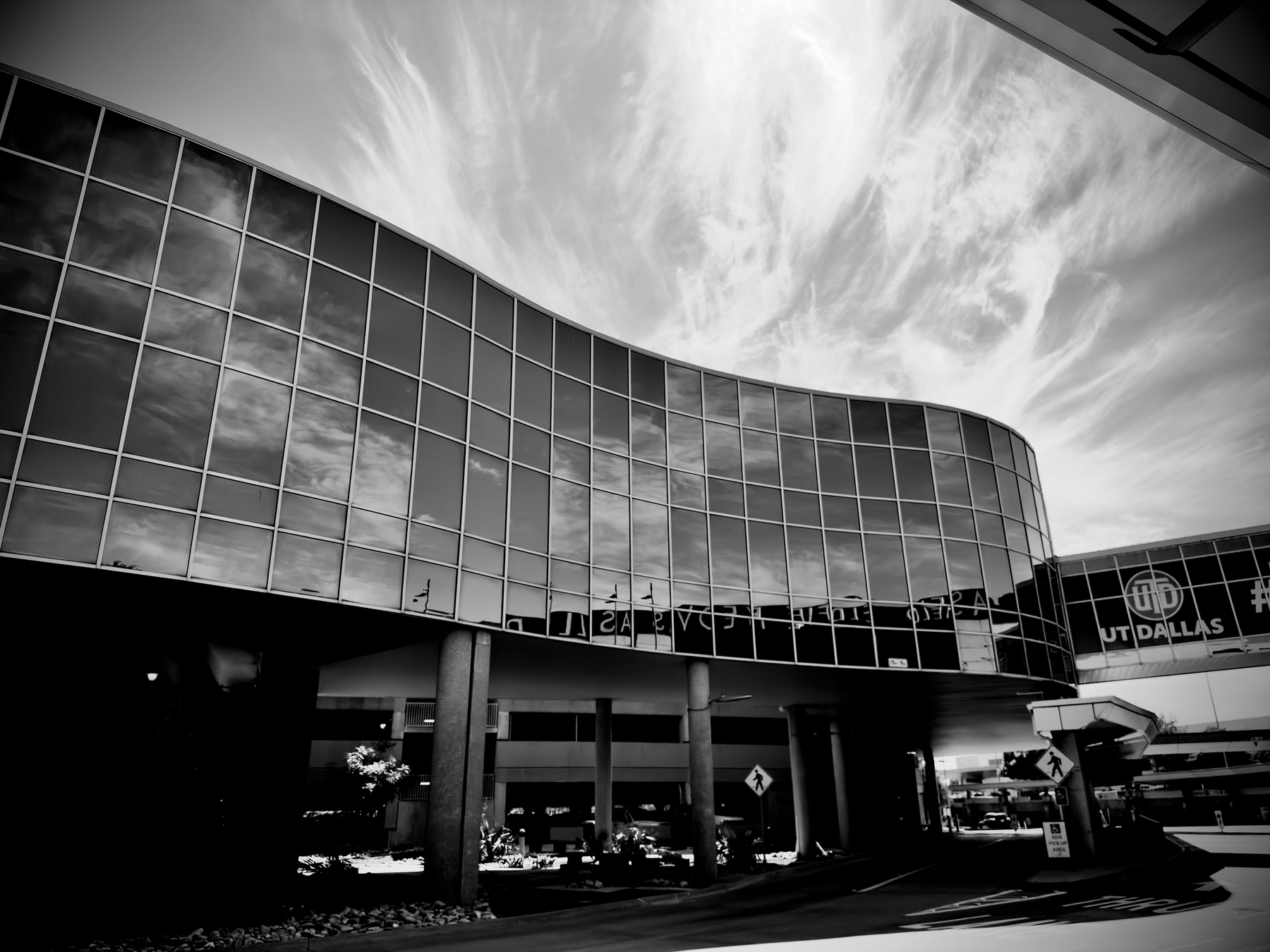 Auto-generated description: A modern building with a wavy glass facade is under a sky filled with wispy clouds, featuring a sign that reads UT DALLAS.