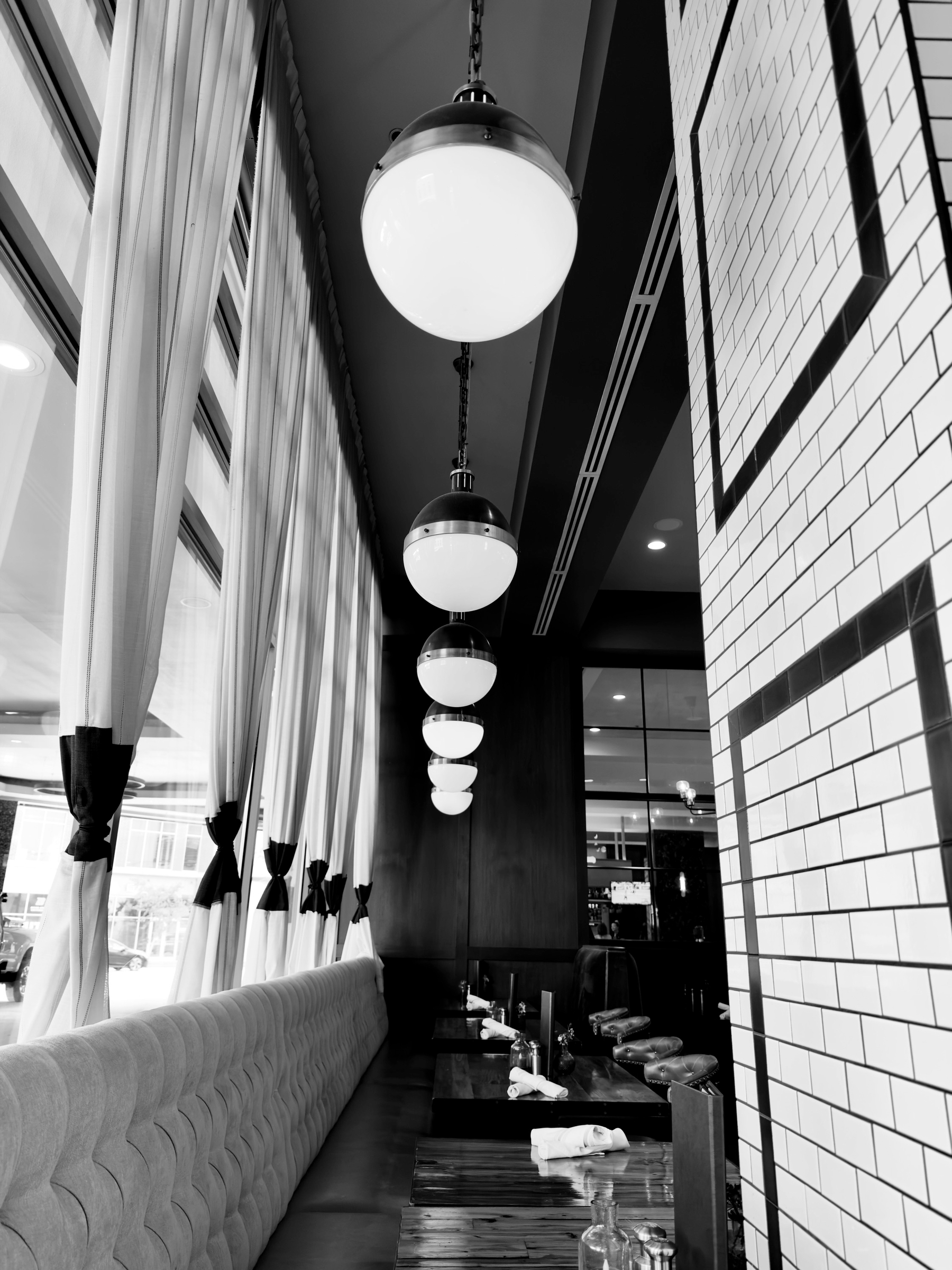 Auto-generated description: A row of spherical pendant lights hangs above a long seating area with tufted upholstery, wooden tables, and large windows with curtains tied back.