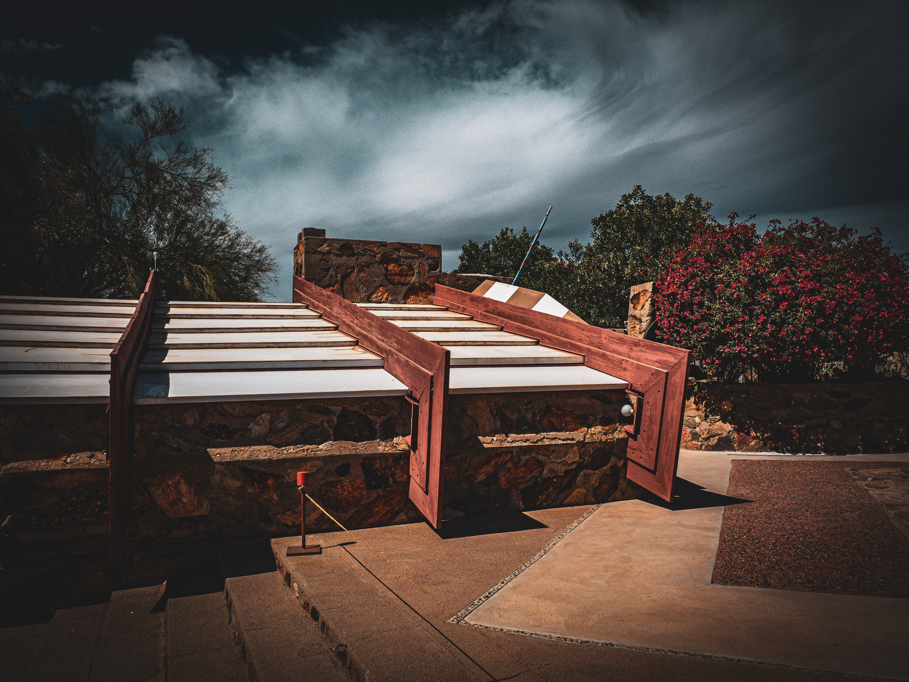Auto-generated description: A modern architectural structure with angled wooden beams and a glass roof is set against a dramatic cloudy sky and surrounded by lush greenery.