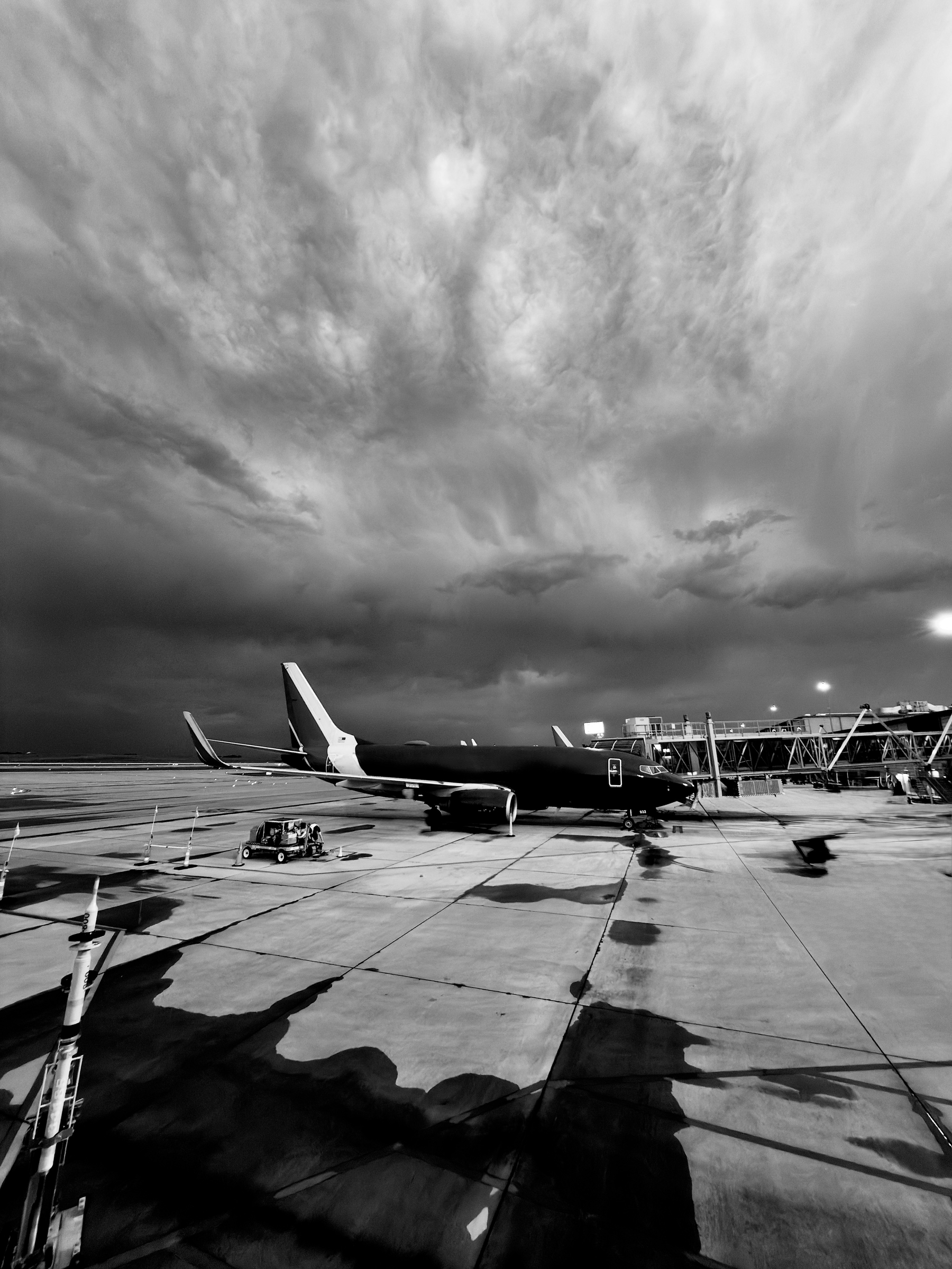 Auto-generated description: A large airplane is parked at the airport gate under a dramatic, cloudy sky in a black and white photo.