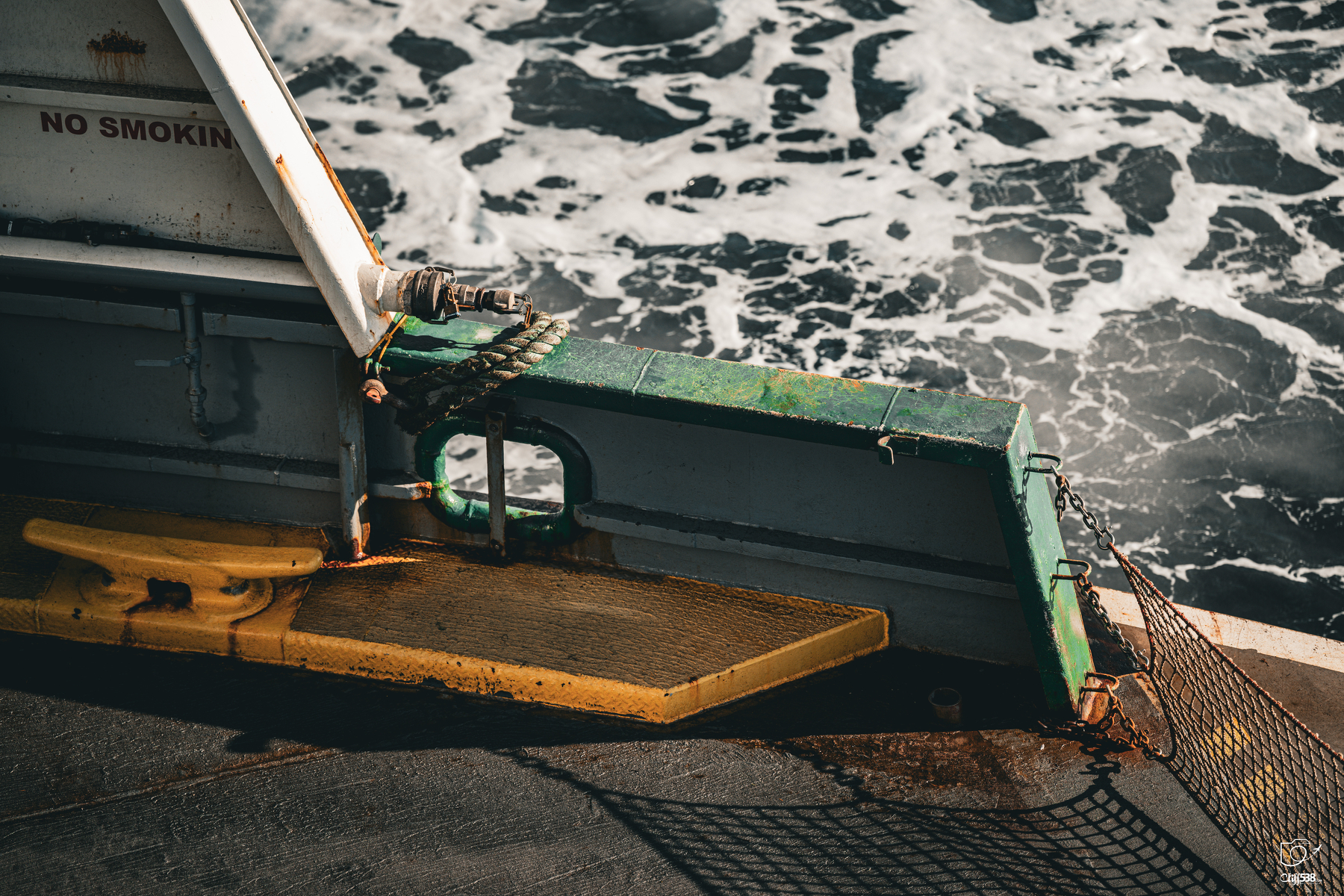 Auto-generated description: Part of a boat deck is seen with a yellow platform, green railing, and churning ocean water in the background.