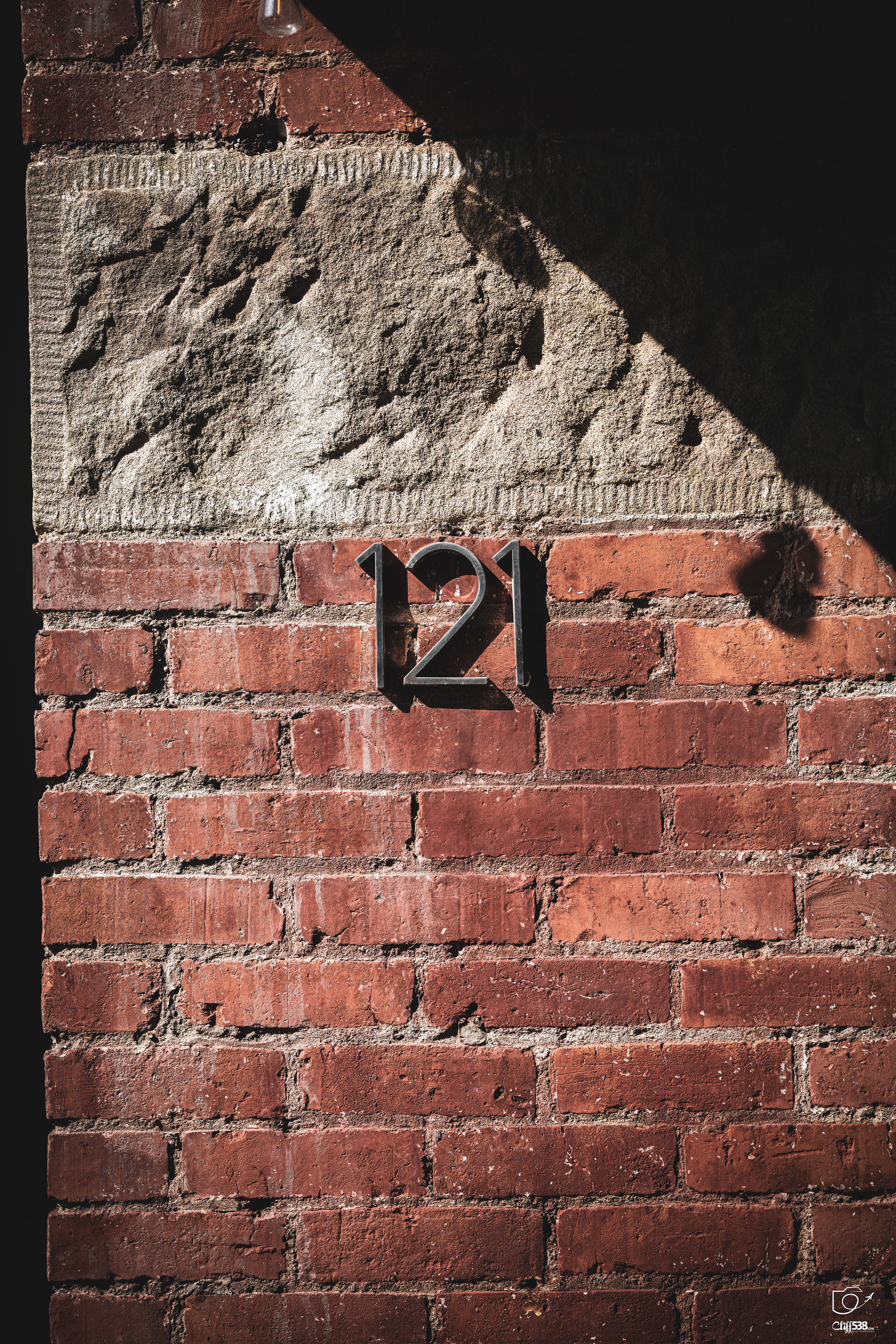 Auto-generated description: The number 121 is displayed on a red brick wall with a rough stone section above it, partially shaded by an overhead structure.