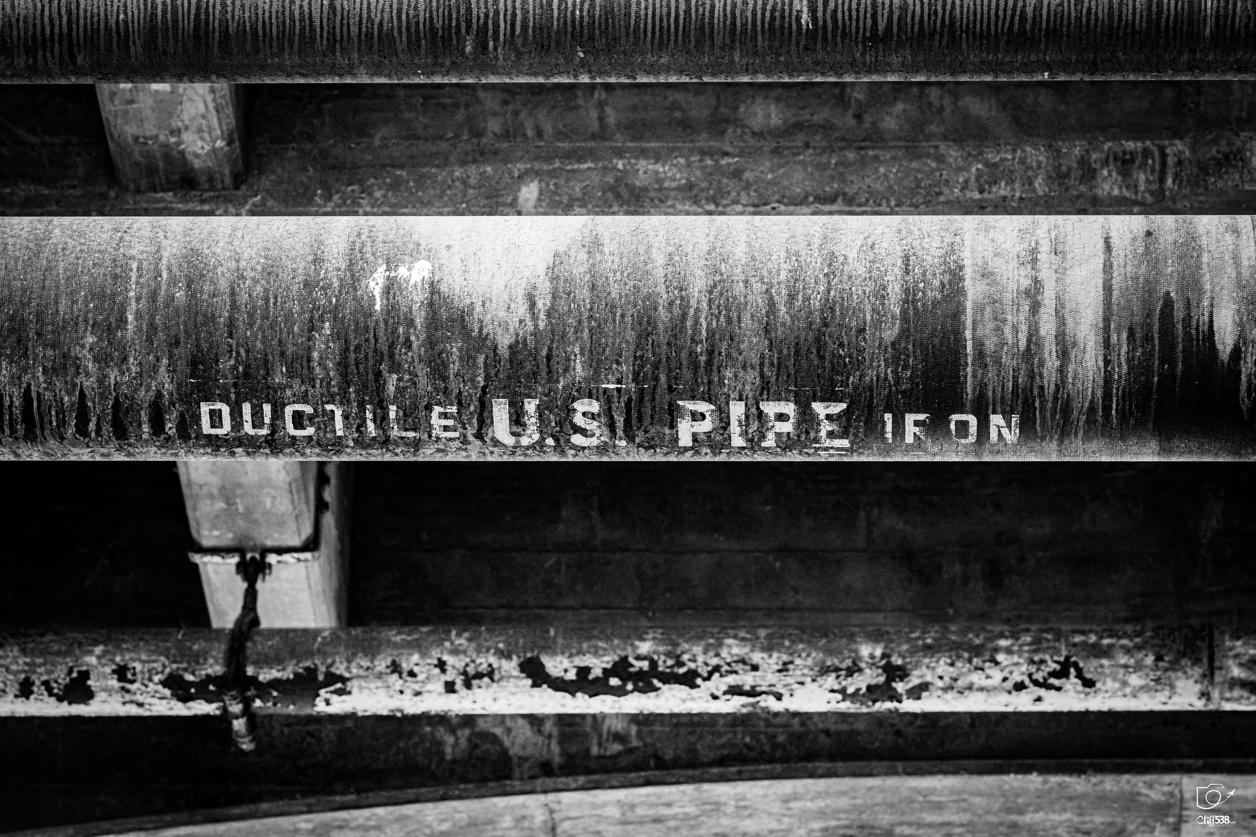 Auto-generated description: A weathered and rusted pipe labeled DUCTILE U.S. PIPE IRON is shown in a black and white photograph.