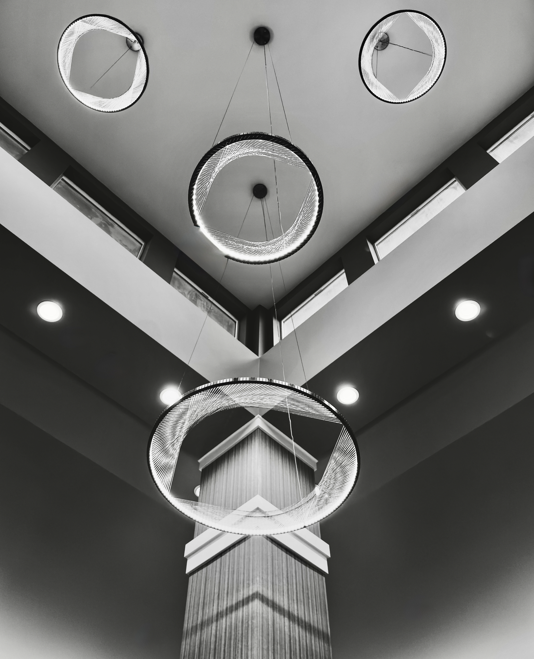 Auto-generated description: A modern architectural ceiling design features circular chandeliers suspended beneath a geometric skylight, enhanced by dramatic contrast and lighting.