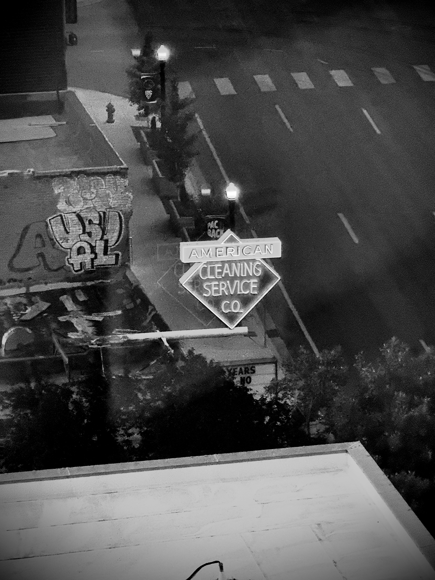 Auto-generated description: A nighttime street scene features a neon sign for American Cleaning Service Co. alongside graffiti on a nearby building and dimly lit street lamps.