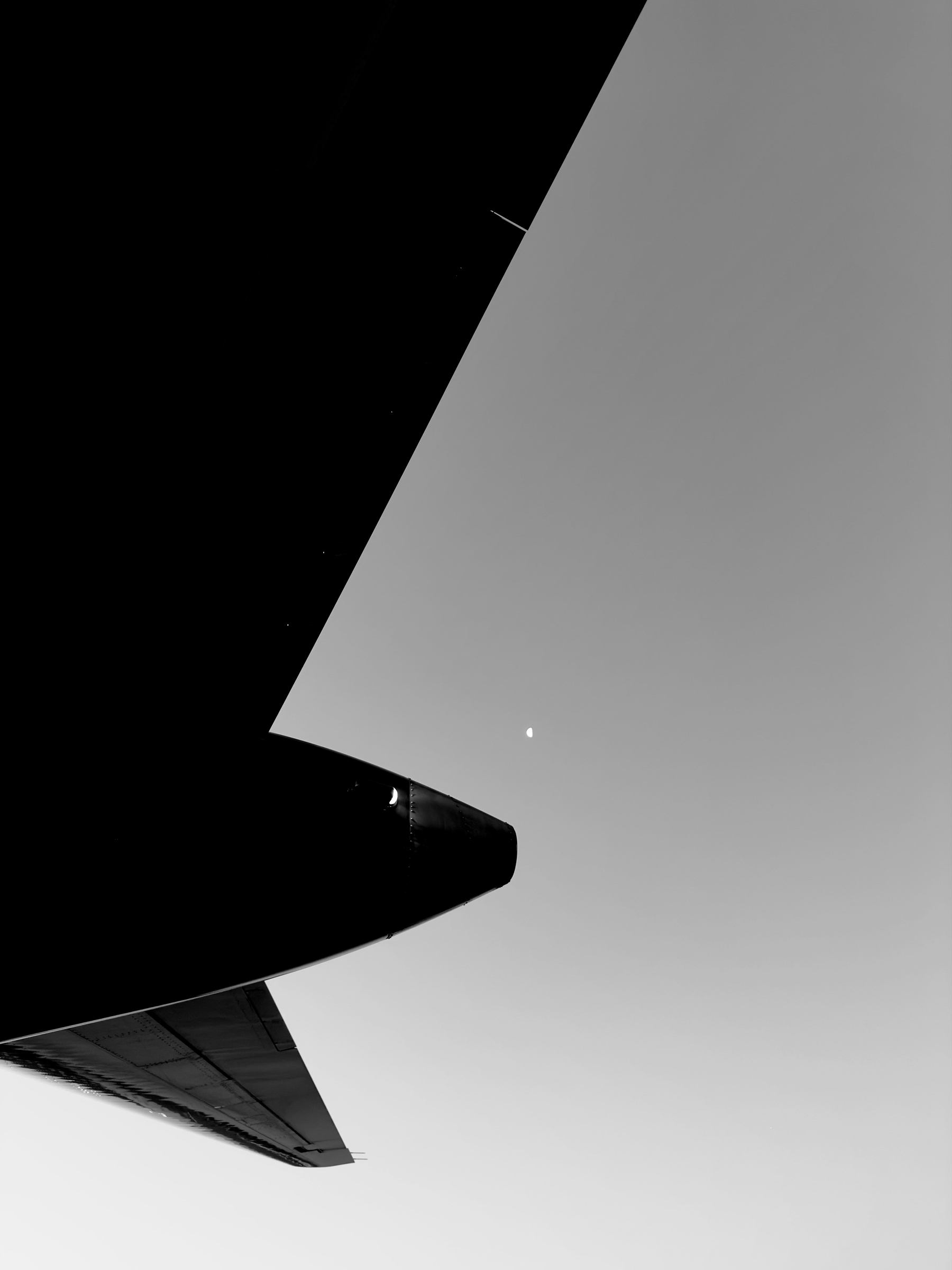 Auto-generated description: A black and white photo capturing an airplane wing with a small bright moon in the clear sky.