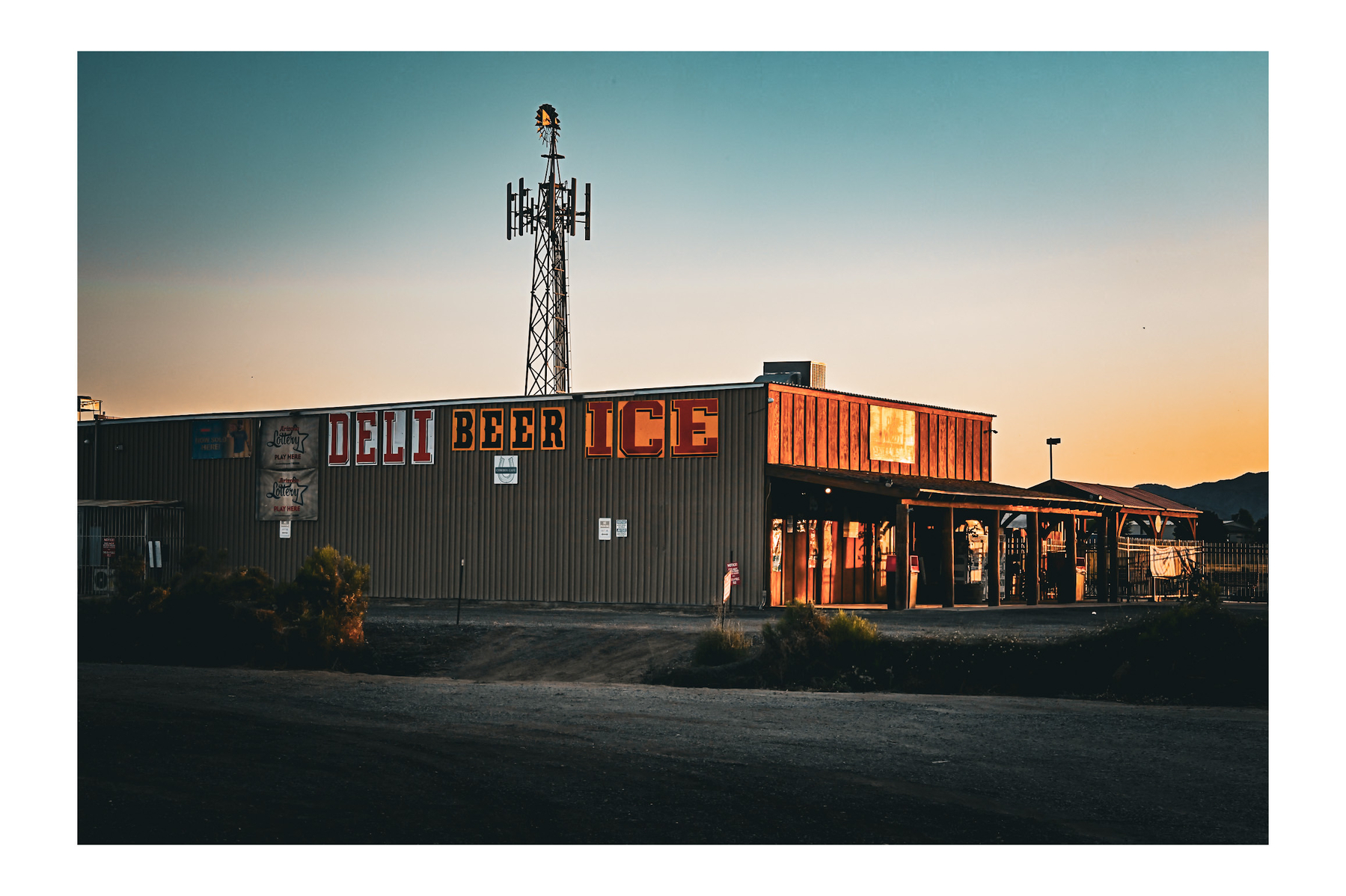 Auto-generated description: A rustic building with signs for deli, beer, and ice stands along a roadside during sunset with a communications tower in the background.