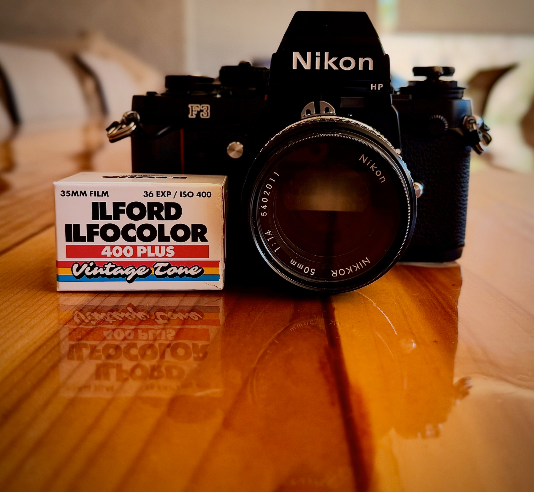 Auto-generated description: A vintage Nikon F3 camera with a Nikkor 50mm lens is placed on a wooden surface next to a box of ILFORD ILFOCOLOR 400 Plus film.