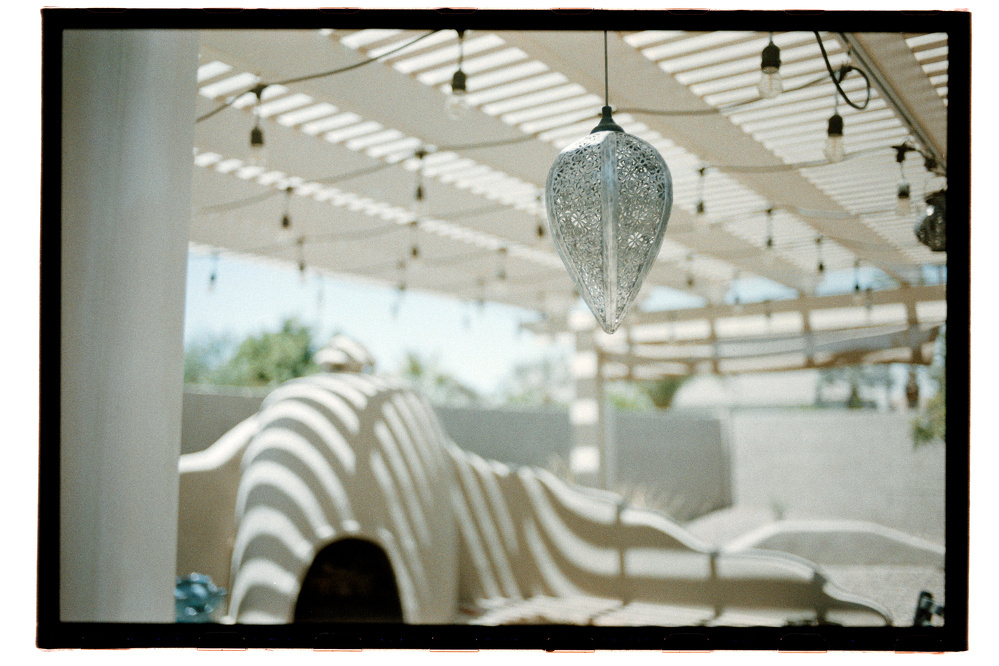 A decorative, intricately patterned pendant light hangs under a pergola with striped shadows cast on curved clay structures.