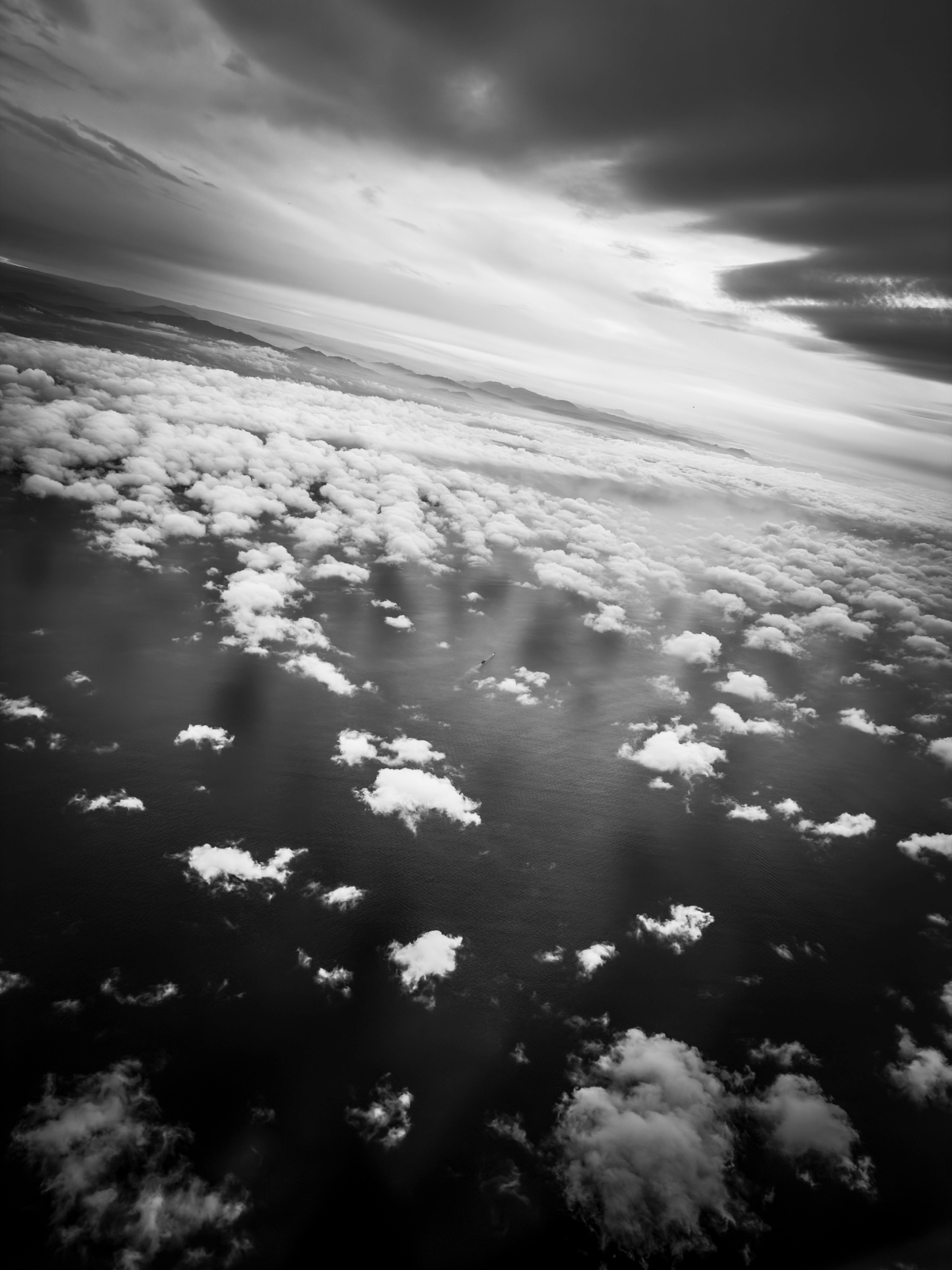 Auto-generated description: A dramatic black and white aerial view captures scattered clouds over a dark landscape beneath a moody, cloud-filled sky.