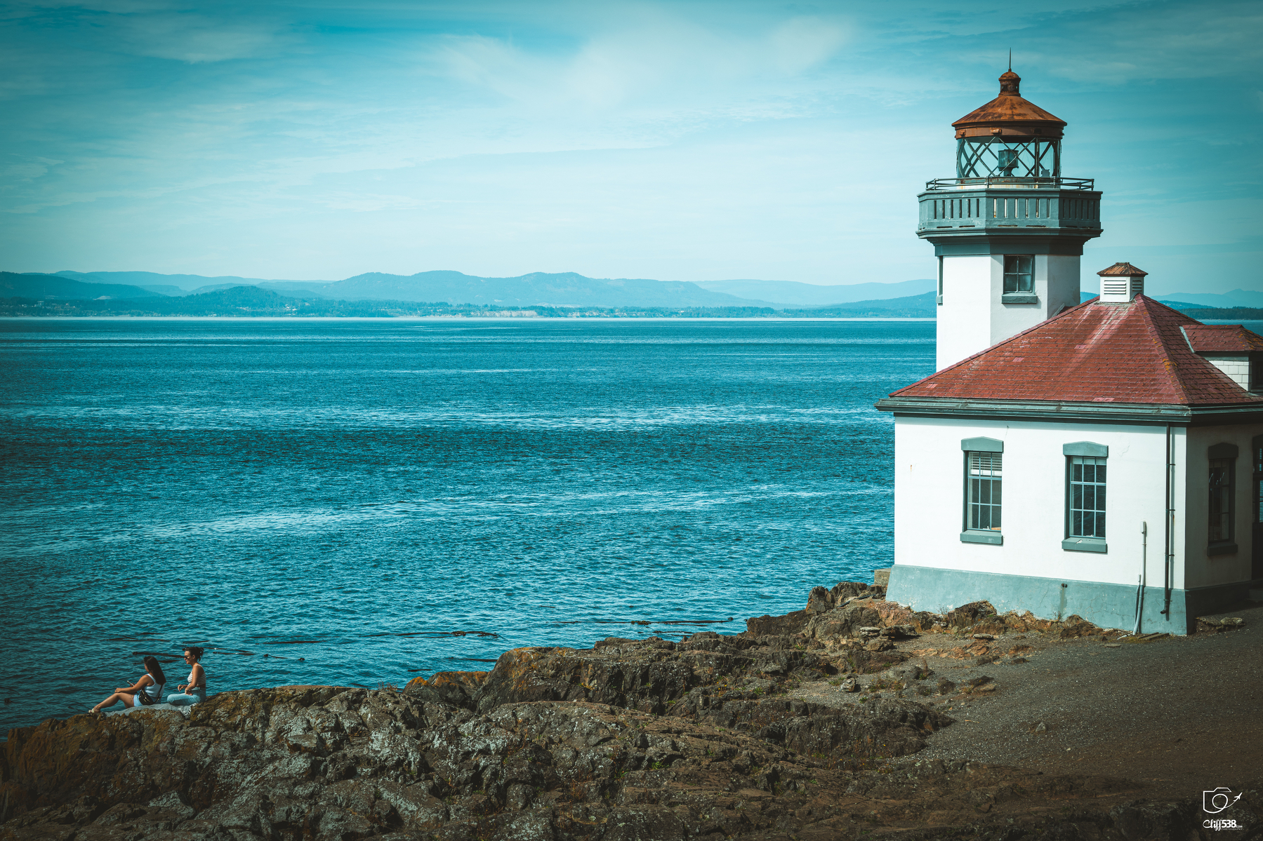 Auto-generated description: A lighthouse stands on a rocky coastline while two people sit near the water's edge overlooking the calm blue sea.