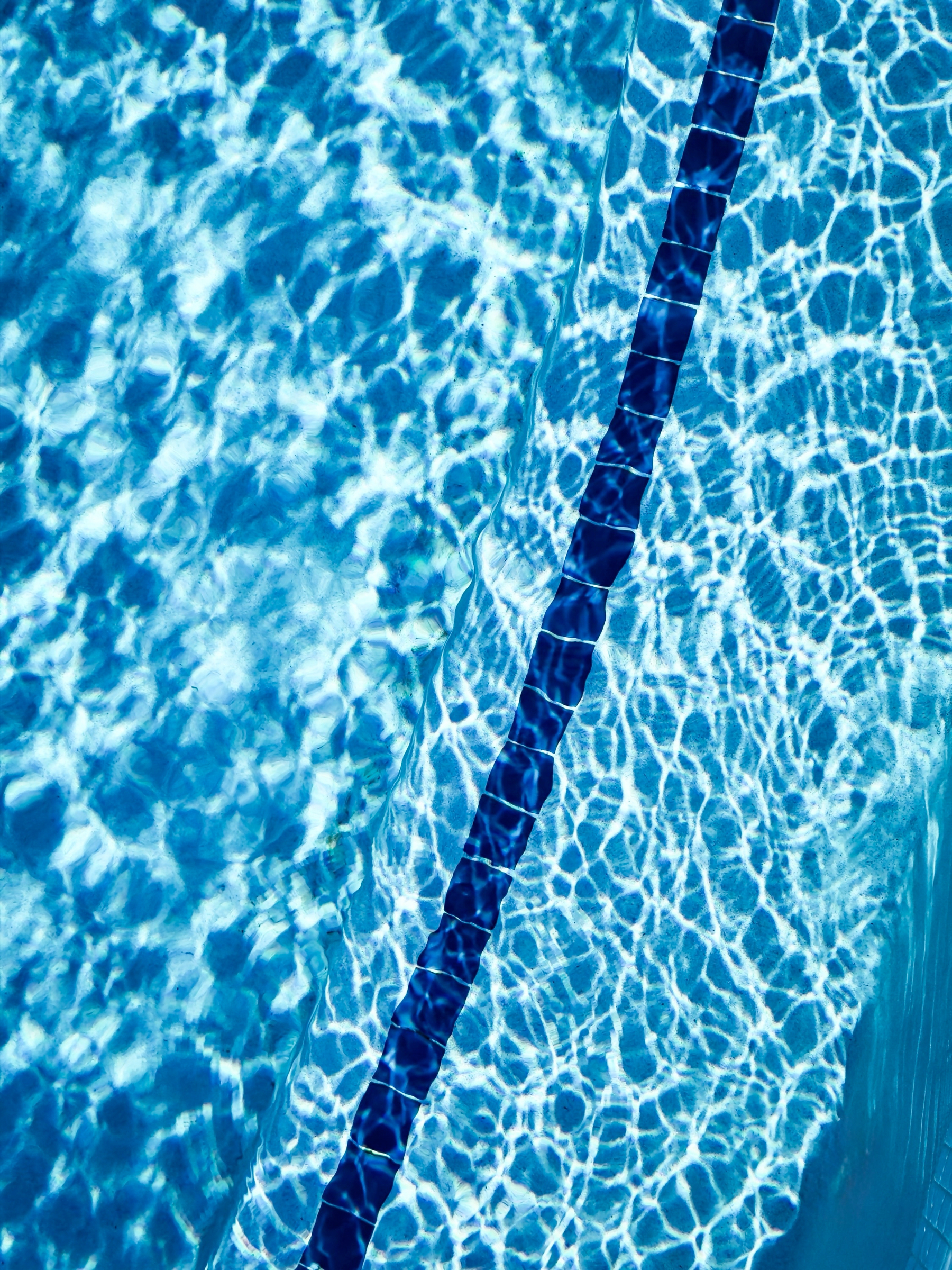 Auto-generated description: Clear blue water with sunlight reflecting on the bottom, separated by a dark pool lane marker.