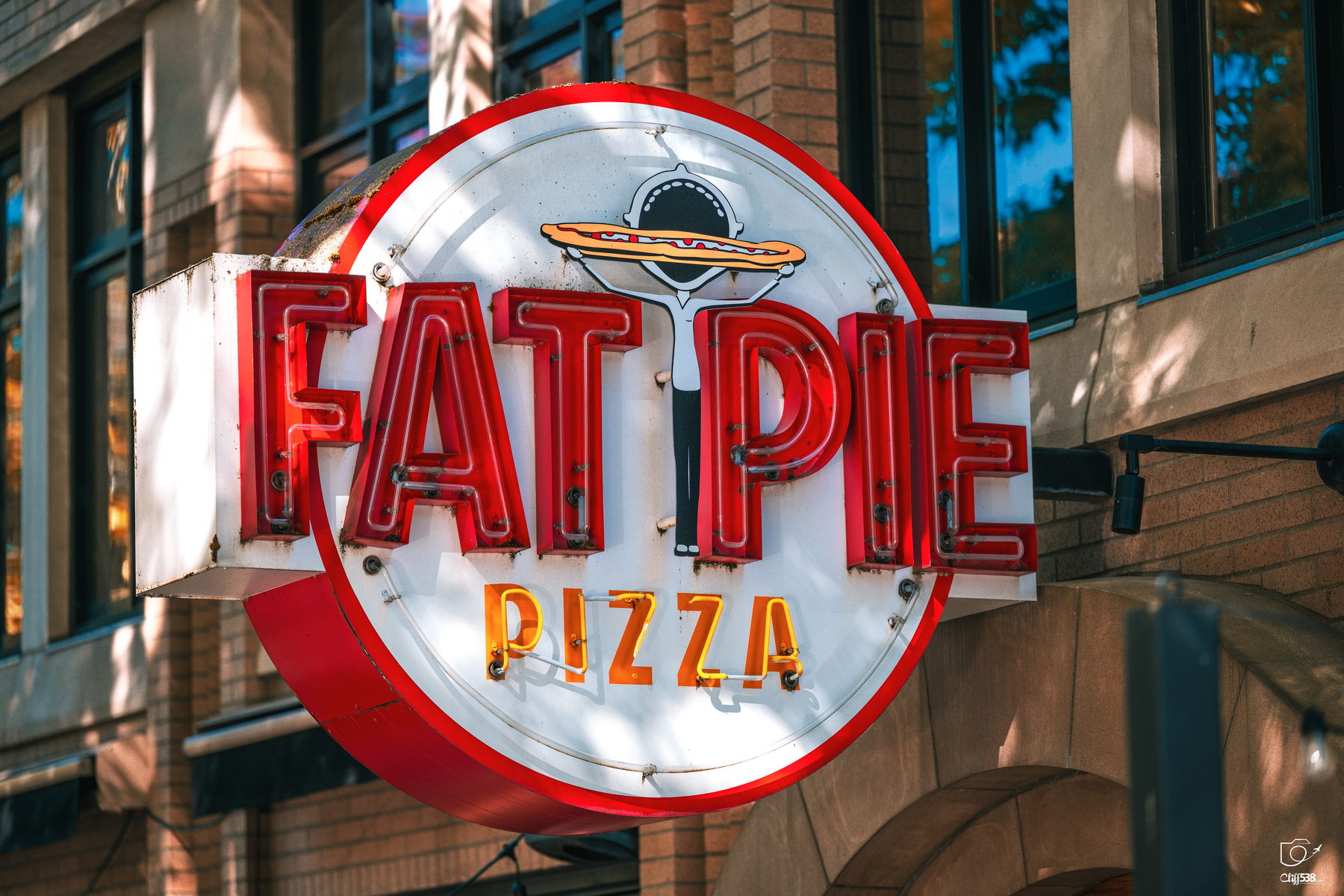 Auto-generated description: A brightly lit neon sign for Fat Pie Pizza is mounted on the side of a brick building.