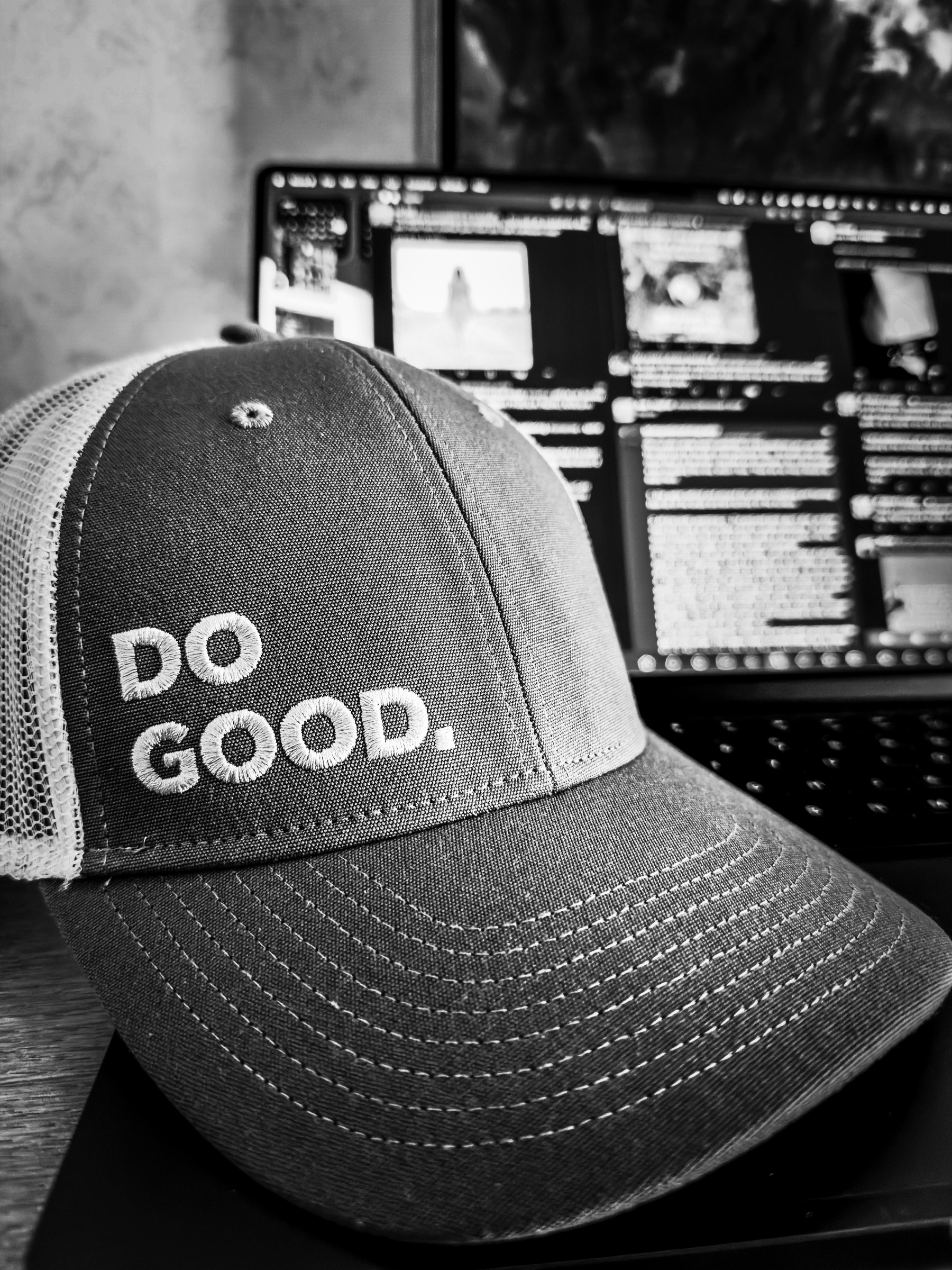 Auto-generated description: A cap with the phrase DO GOOD. embroidered on it rests in front of a laptop displaying various web pages.
