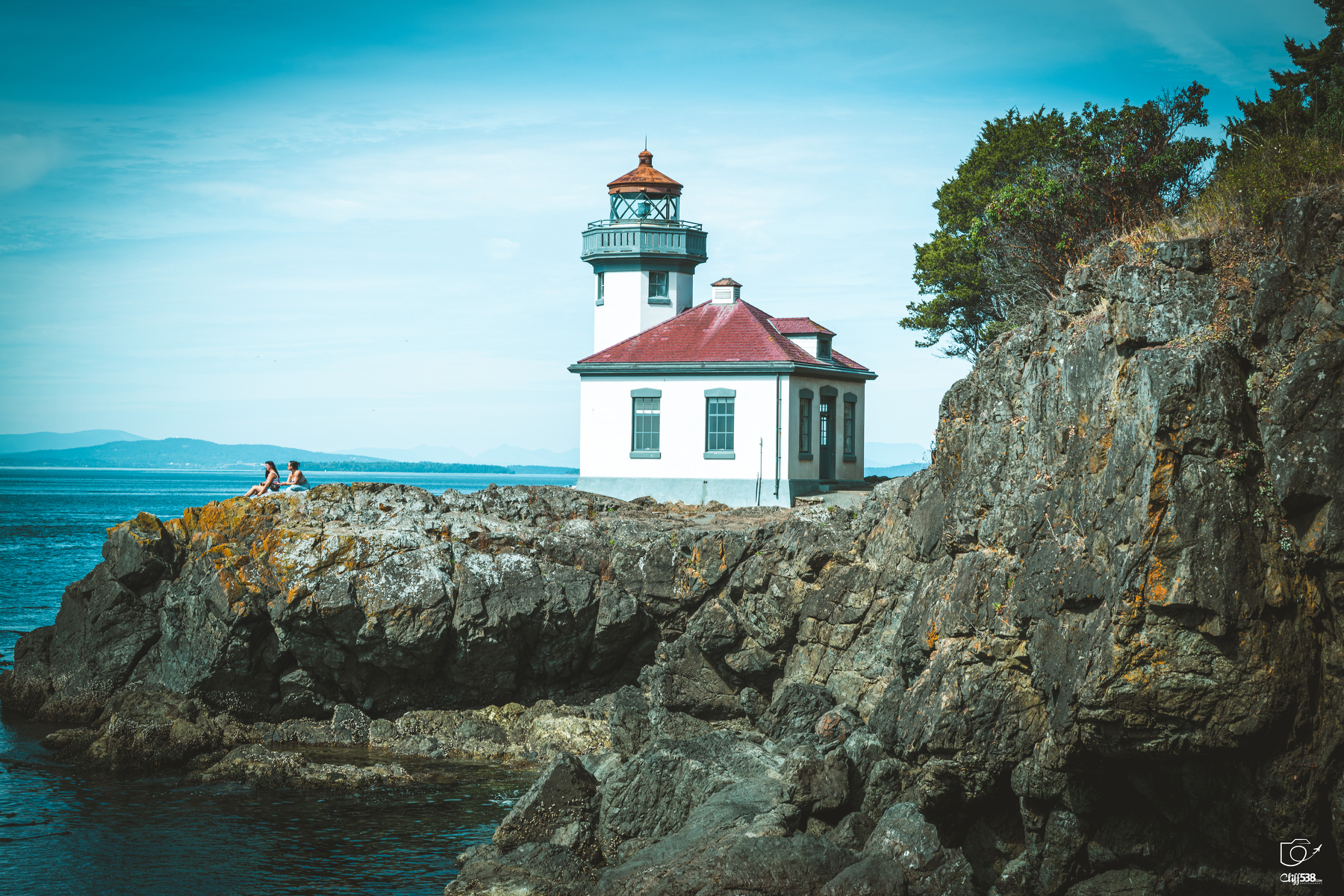 Auto-generated description: A lighthouse with a red roof stands on rugged cliffs by the sea under a clear blue sky.