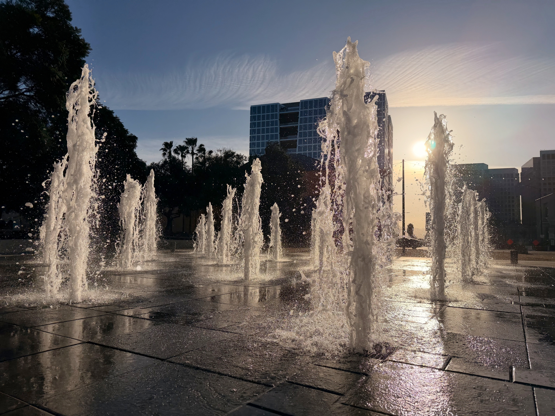 Auto-generated description: Fountains spray water into the air against a backdrop of a sunset and city buildings.