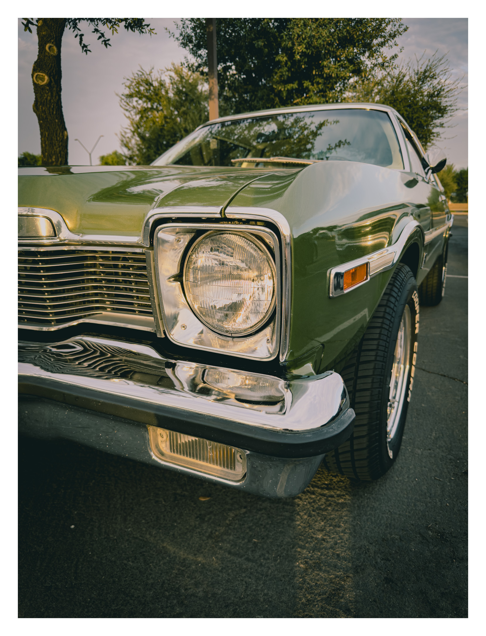A close-up view of the front side of a green classic car parked on a street with trees in the background.
