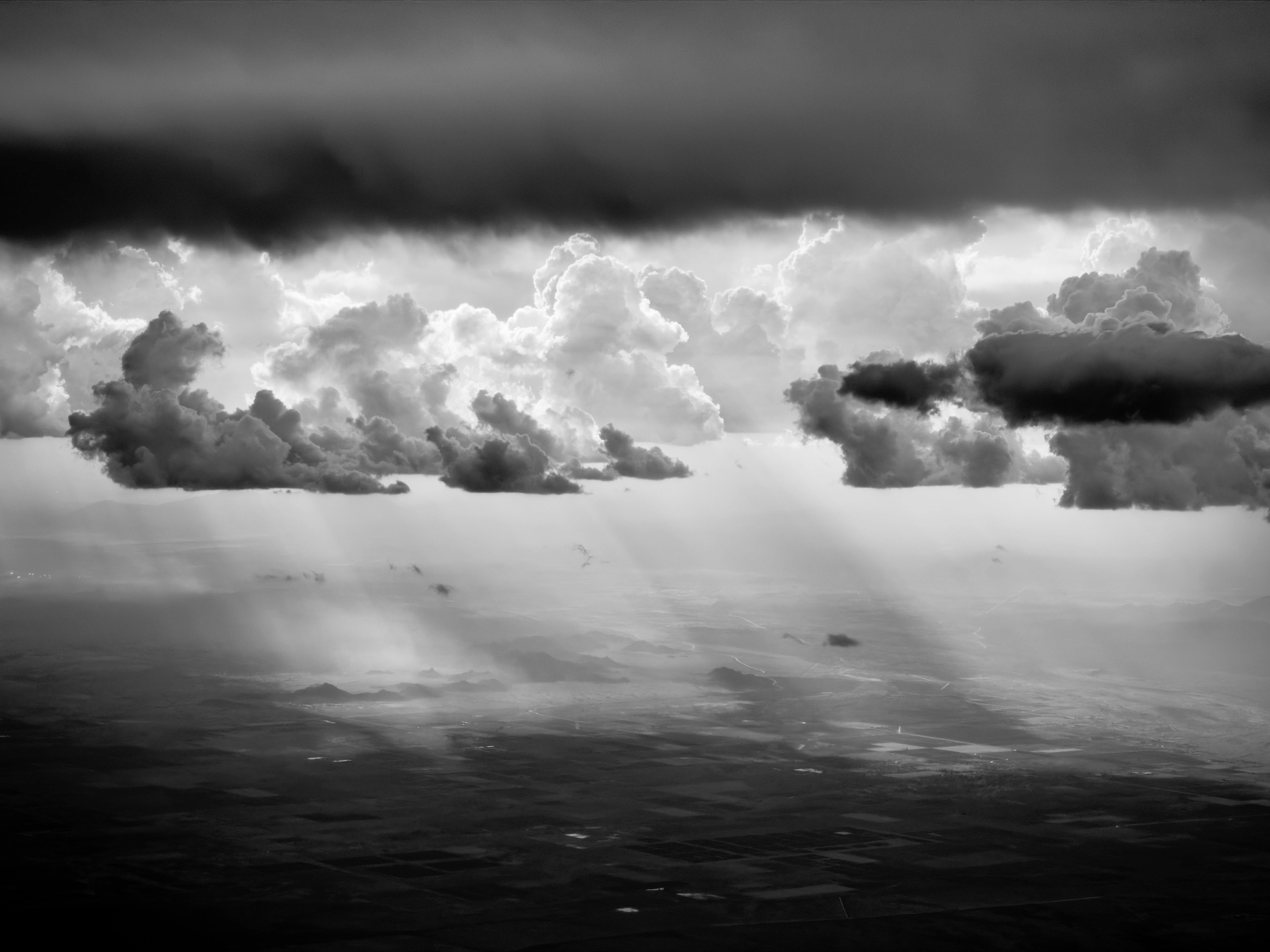 Auto-generated description: Dramatic clouds cast sun rays over a vast, shadowed landscape in a black-and-white scene.