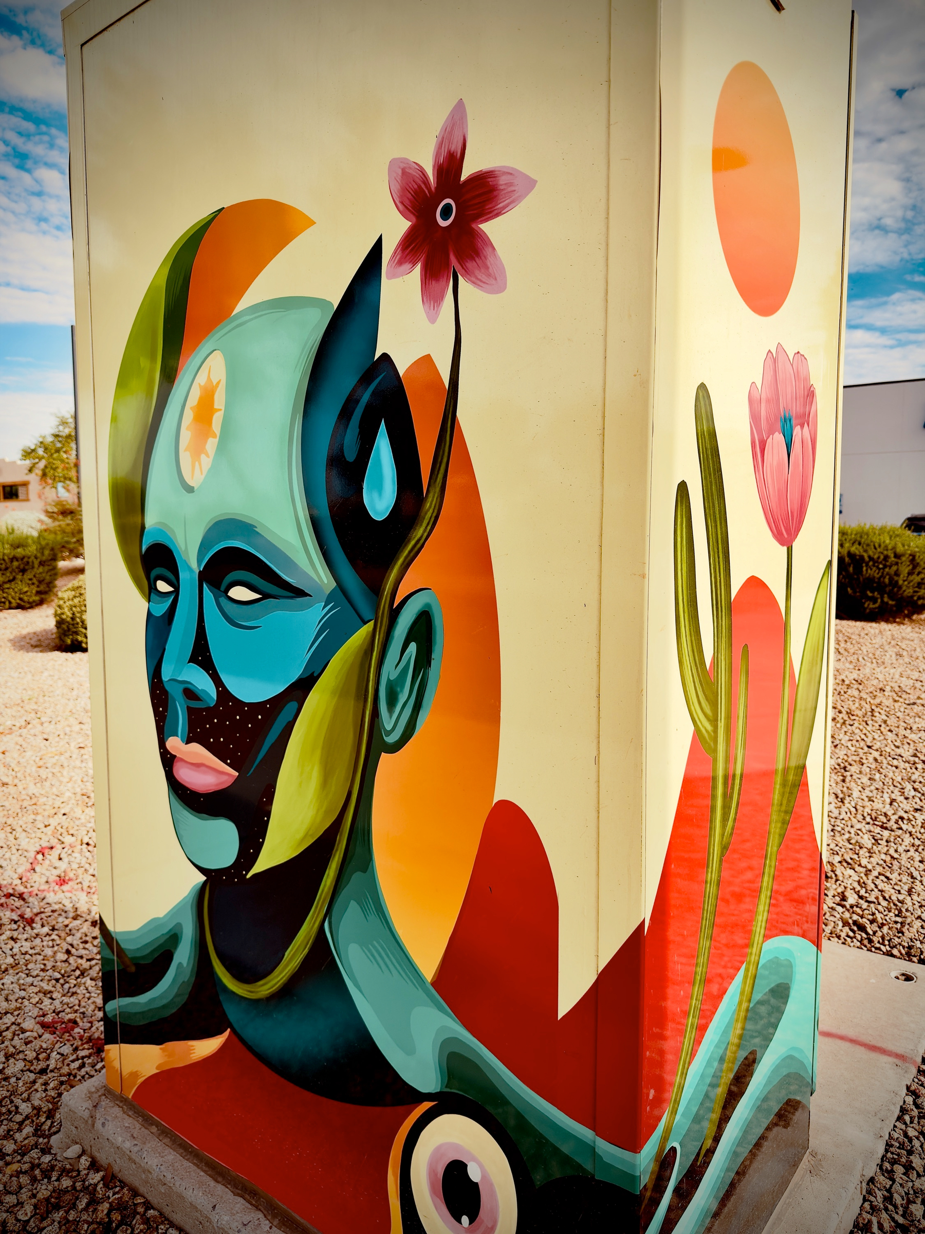 Auto-generated description: A colorful mural on a utility box depicts a stylized blue-skinned figure with plant-like features, including flowers growing from the head, against an abstract background.