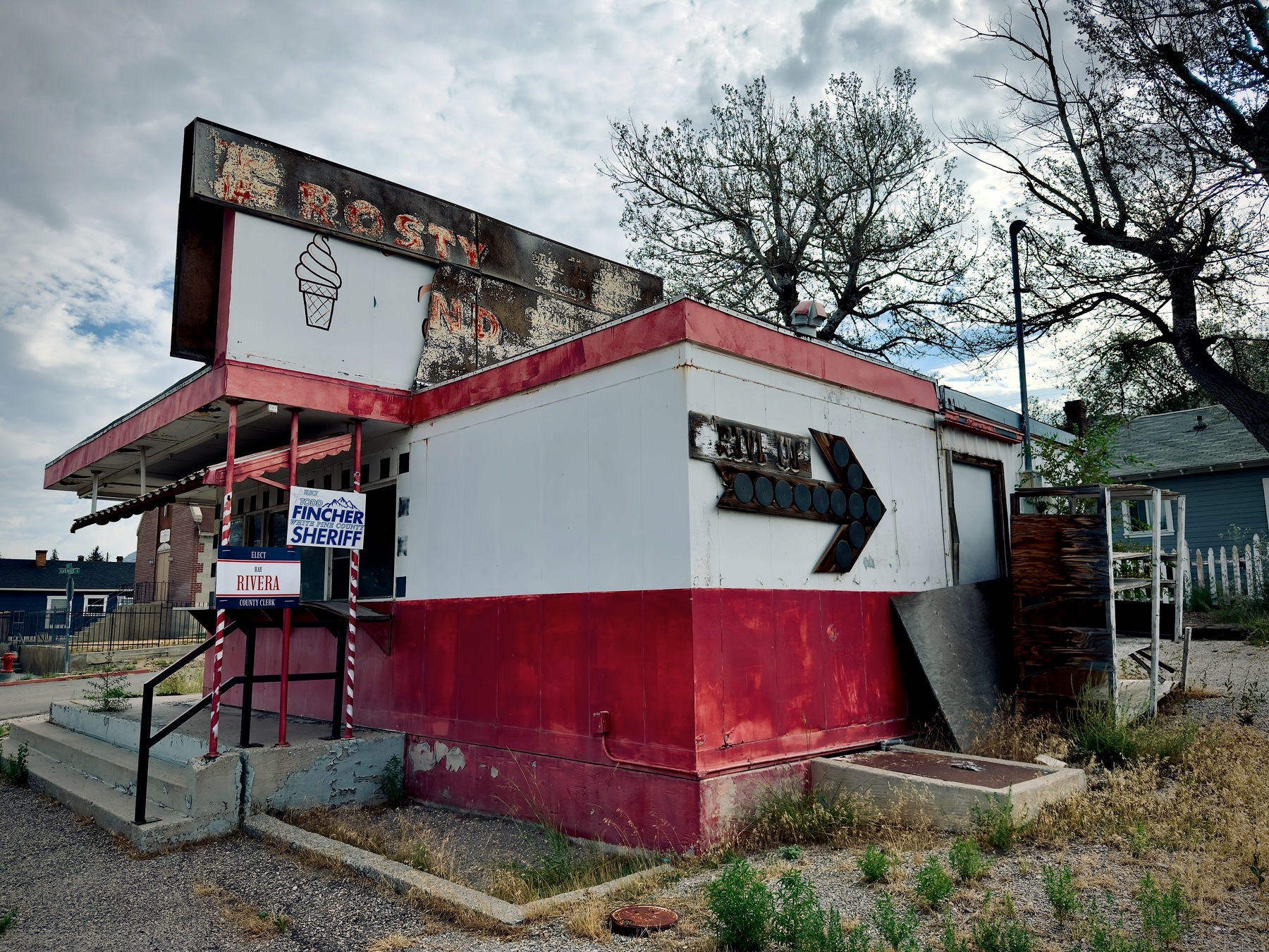 Auto-generated description: A small, weathered building with faded signage, including an ice cream cone illustration and political campaign posters for sheriff and other candidates, stands in a dry, overgrown area.