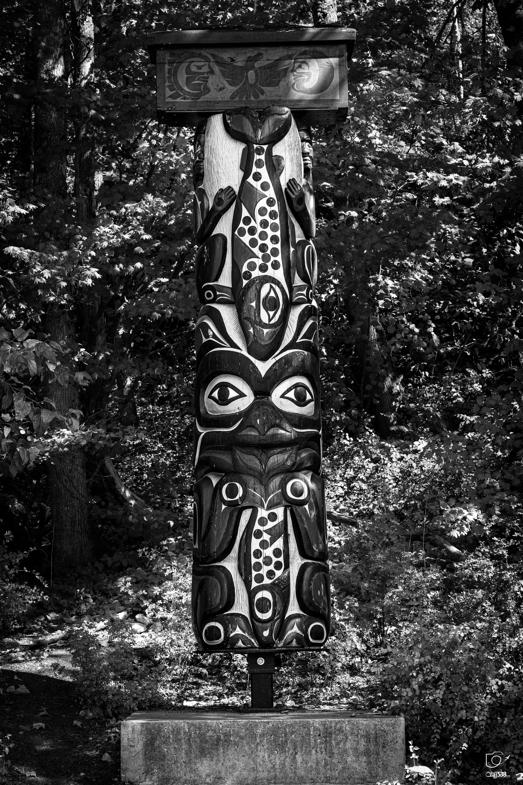 Auto-generated description: A totem pole featuring traditional indigenous artwork stands in a forested area on a concrete base.
