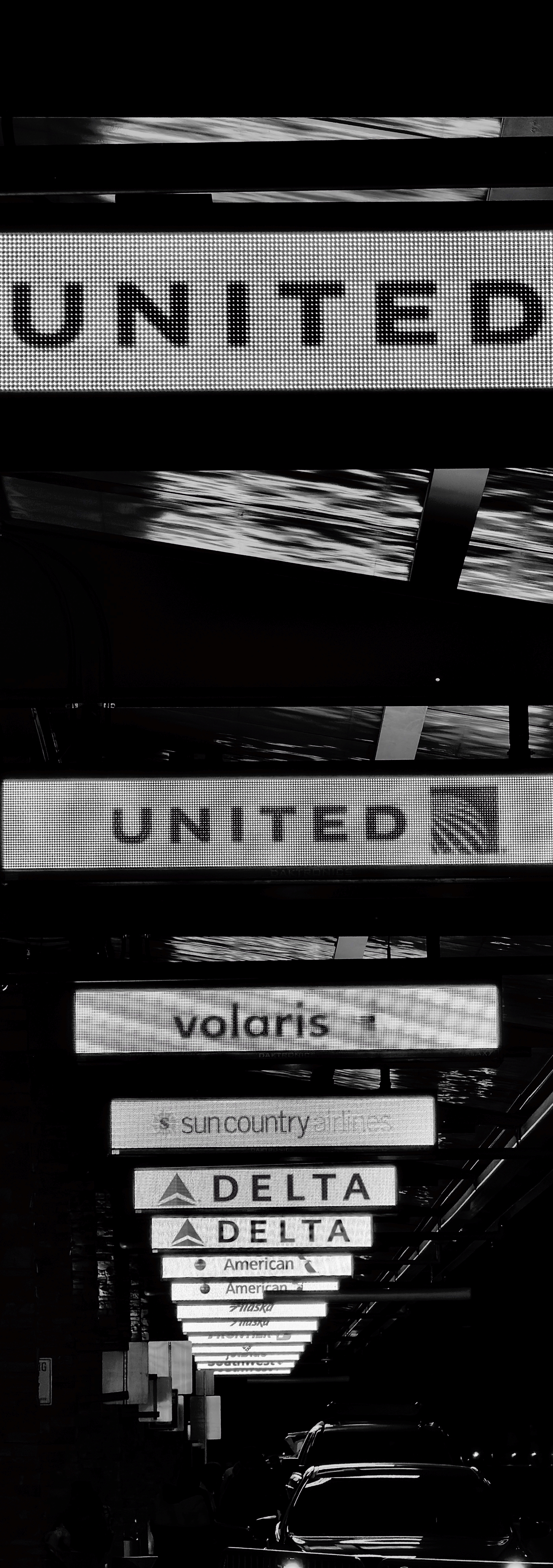Auto-generated description: Multiple airport airline signs including United, Volaris, Sun Country Airlines, Delta, and American are displayed above a roadway with cars beneath them.