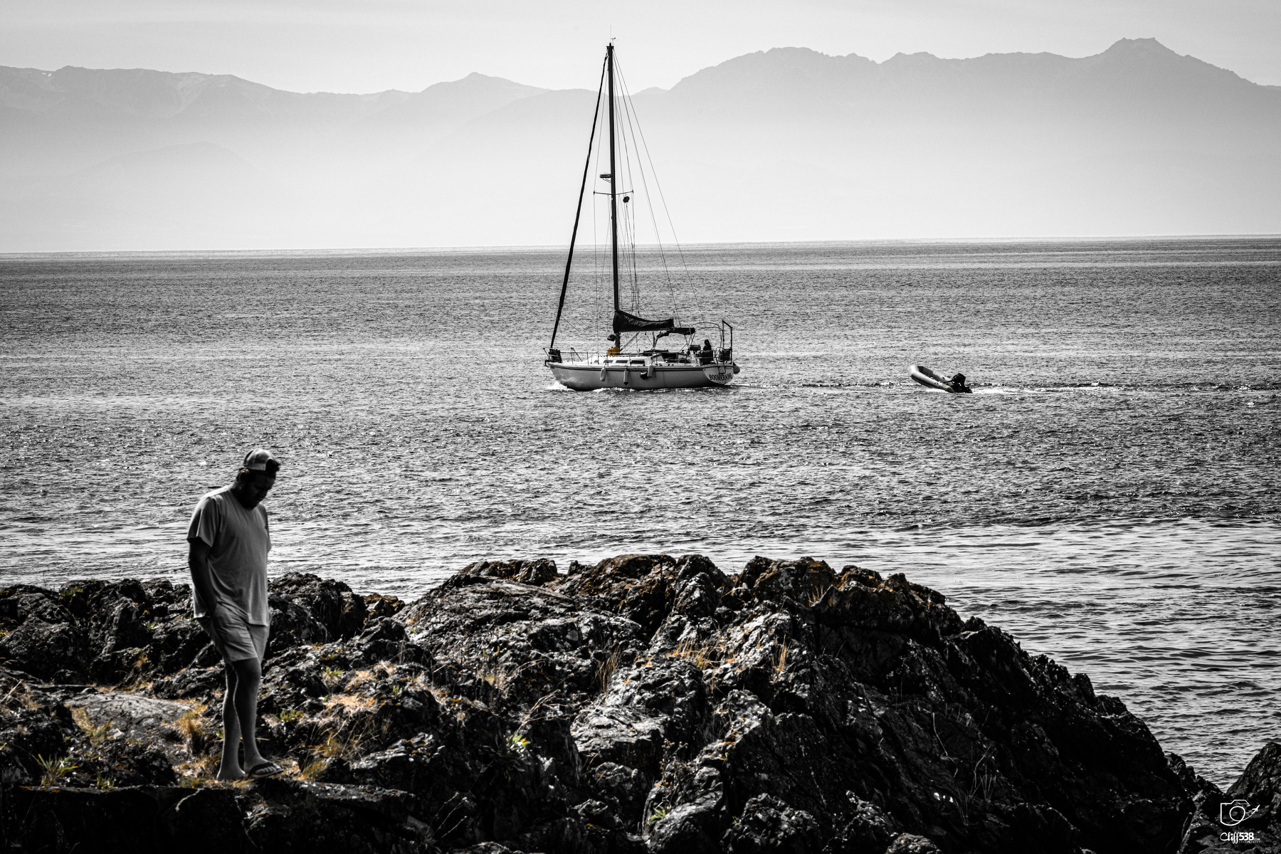 Auto-generated description: A person stands on rocky terrain near the shore while a sailboat floats on the calm sea with mountains visible in the distance.