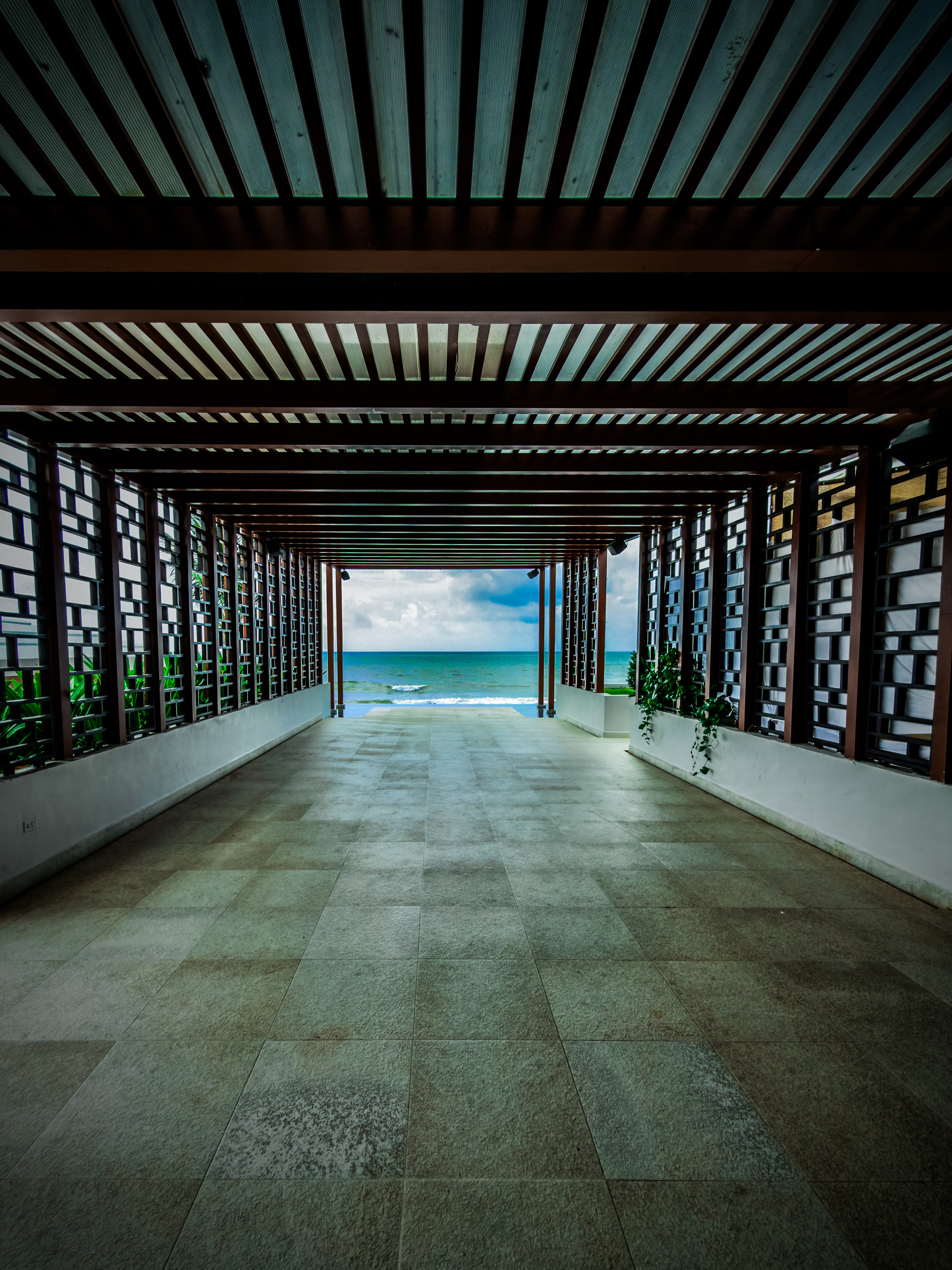 A covered walkway with wooden slats leads to a view of the ocean under a cloudy sky.