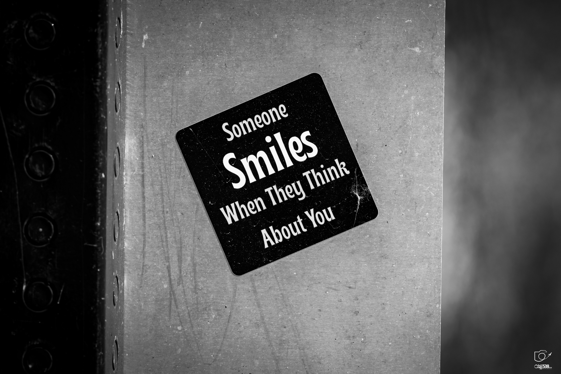 Auto-generated description: A black sticker with white text saying Someone Smiles When They Think About You is attached to a gray, textured surface.