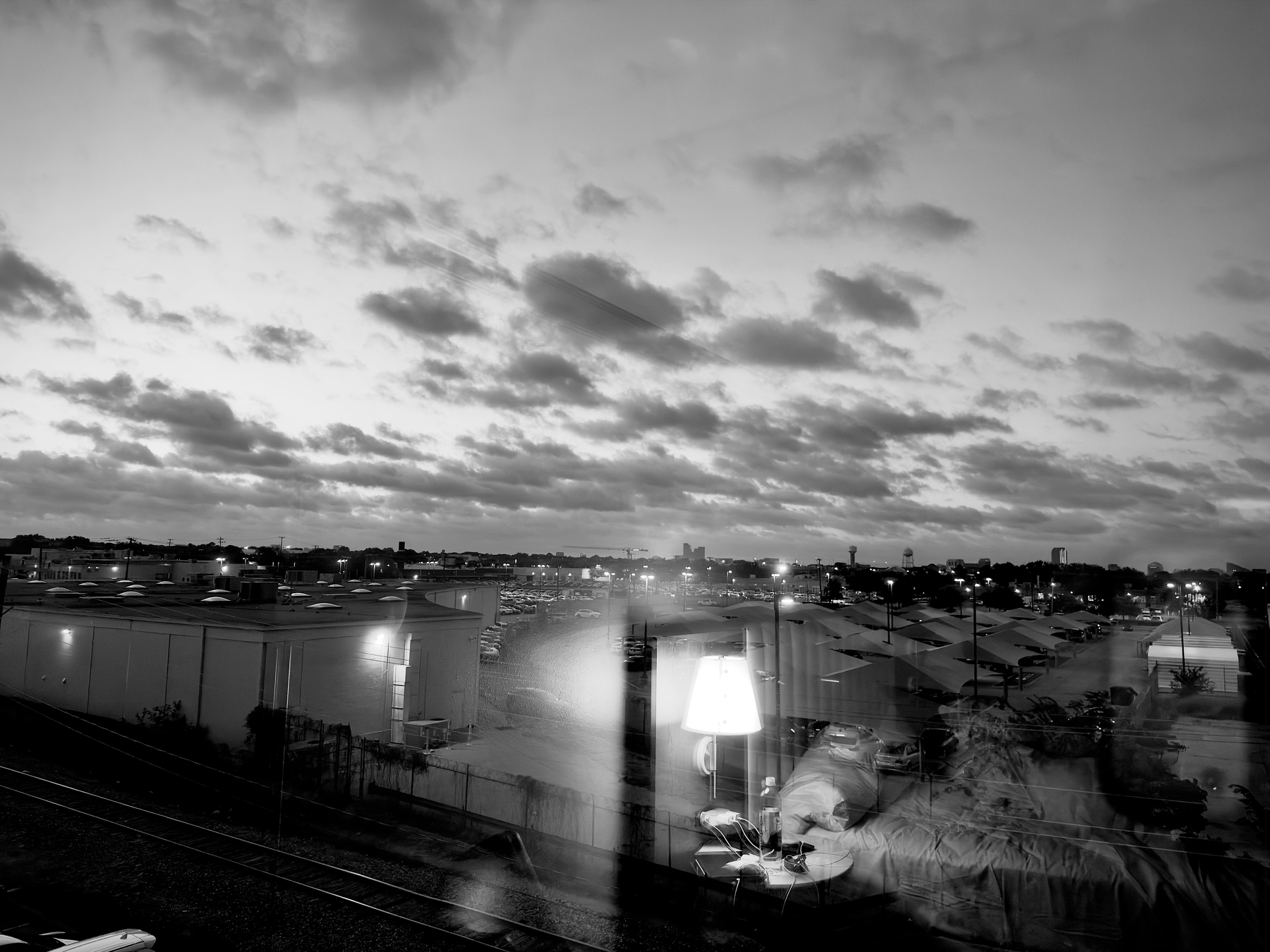 Auto-generated description: A cityscape at dusk is seen through a window reflecting an interior scene with a lit lamp, furniture, and a person.
