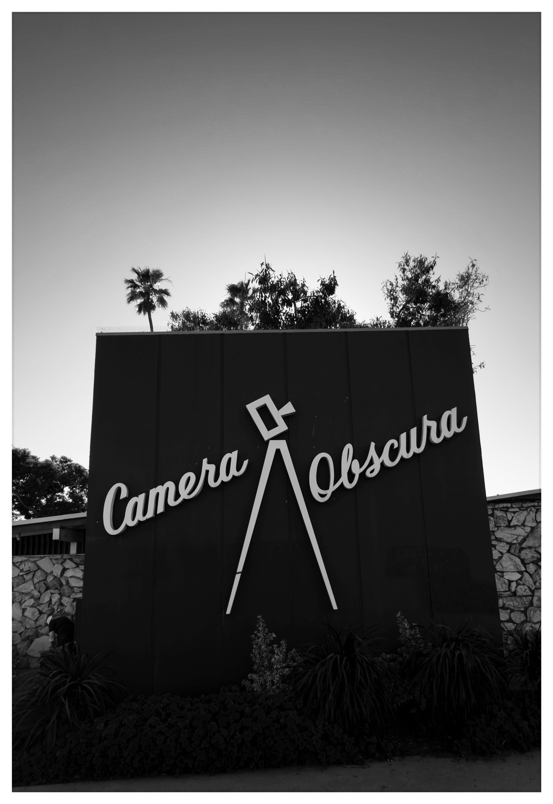 Auto-generated description: A large sign with the words Camera Obscura stands against a backdrop of trees and a clear sky.