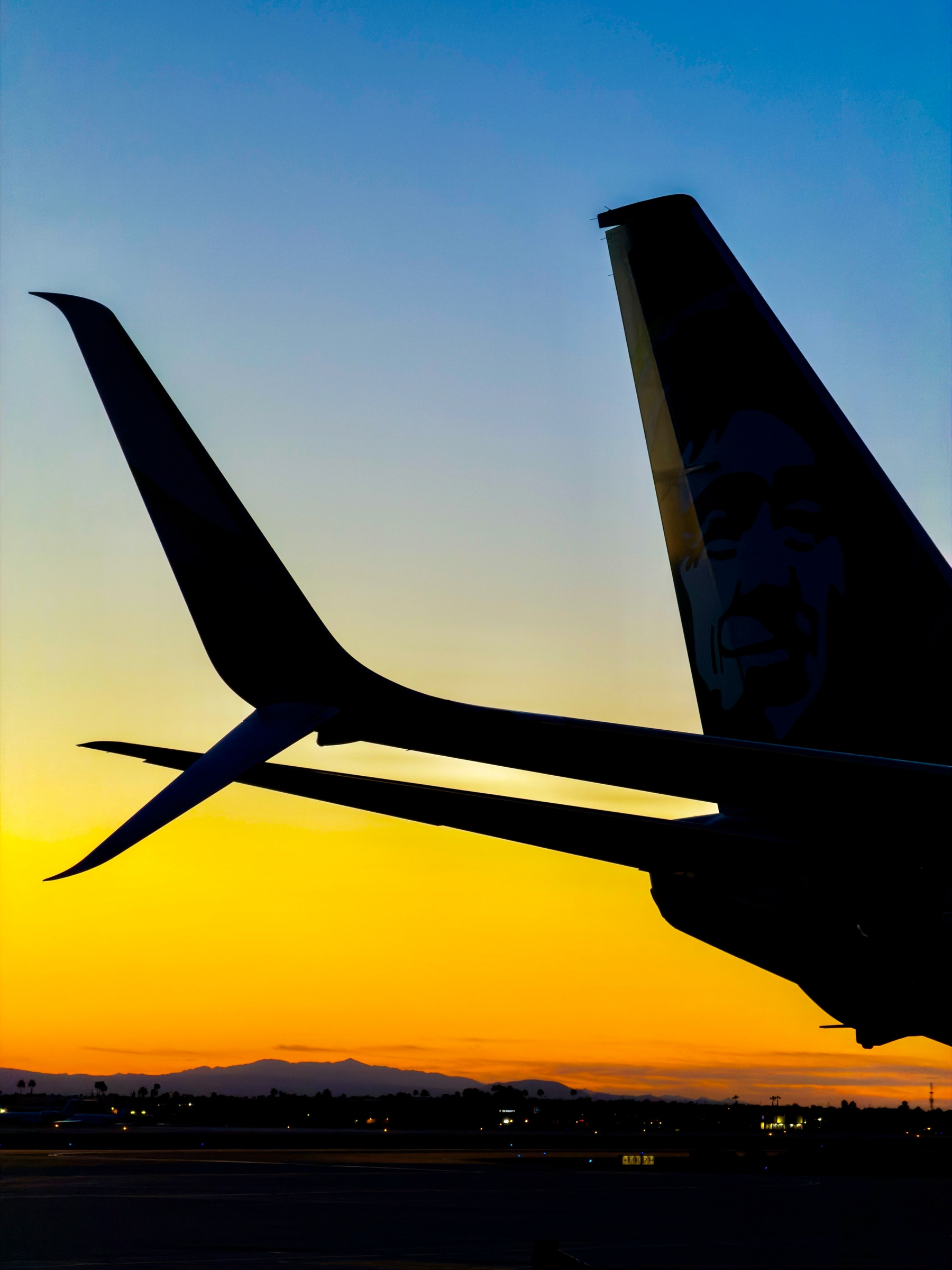 Auto-generated description: A silhouetted airplane tail is set against a vibrant sunset sky with mountains in the distance.