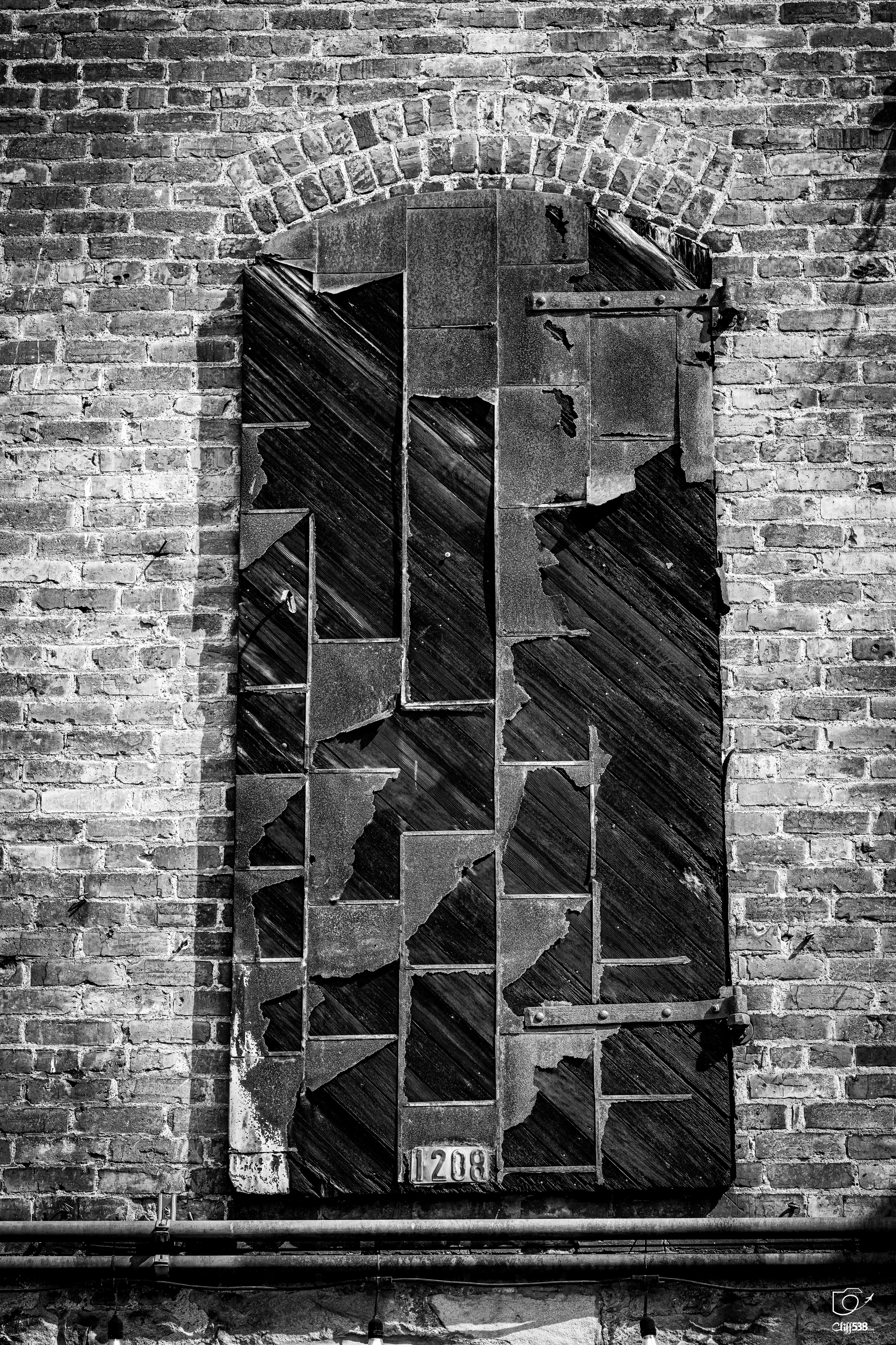 Auto-generated description: A worn, patchwork wooden door with peeling paint is set within an arched brick wall.