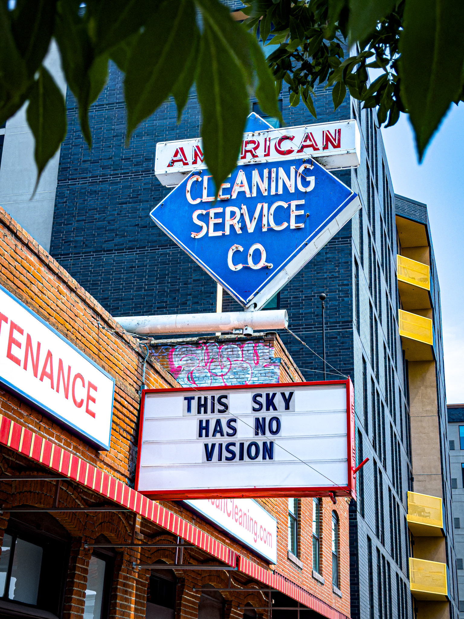 Auto-generated description: A vintage sign for American Cleaning Service Co. is displayed above a marquee with the phrase THIS SKY HAS NO VISION against a backdrop of urban buildings and leafy branches.