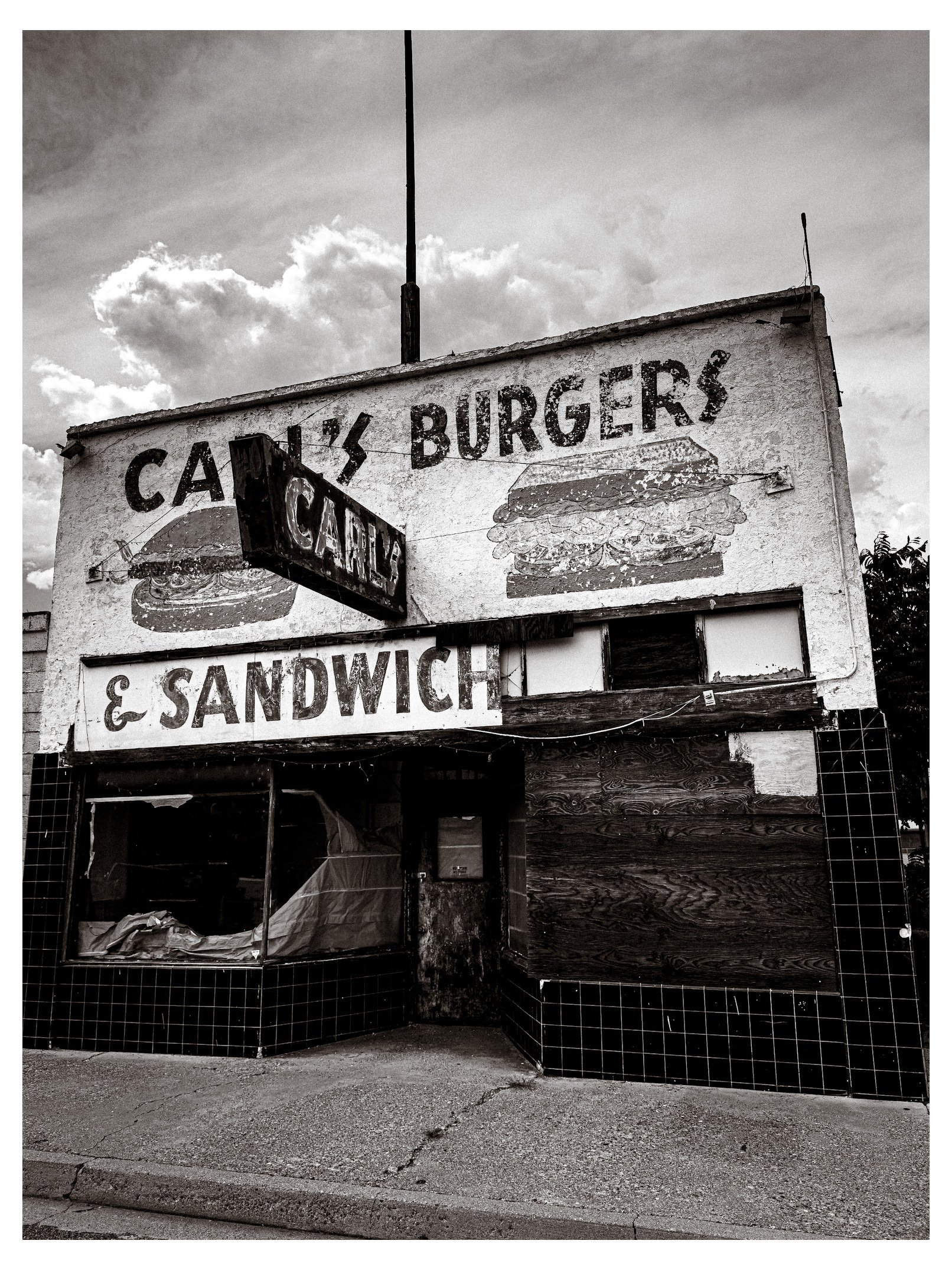 Auto-generated description: An old, weathered storefront advertises Carl's Burgers & Sandwiches with faded signage and boarded-up windows under a cloudy sky.