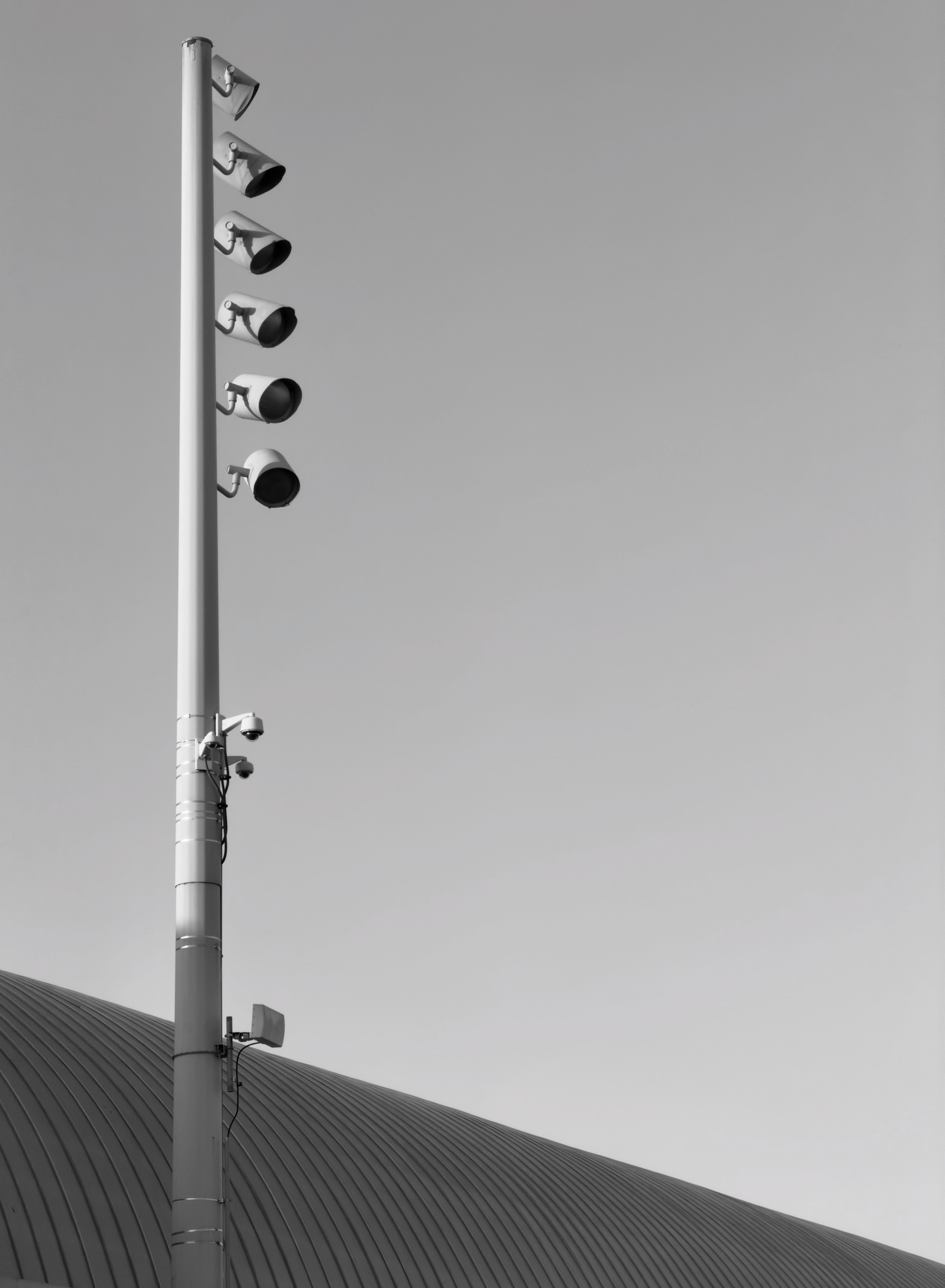 Auto-generated description: A tall pole equipped with several large spotlights and surveillance cameras stands next to a curved, ribbed roof under a clear sky.