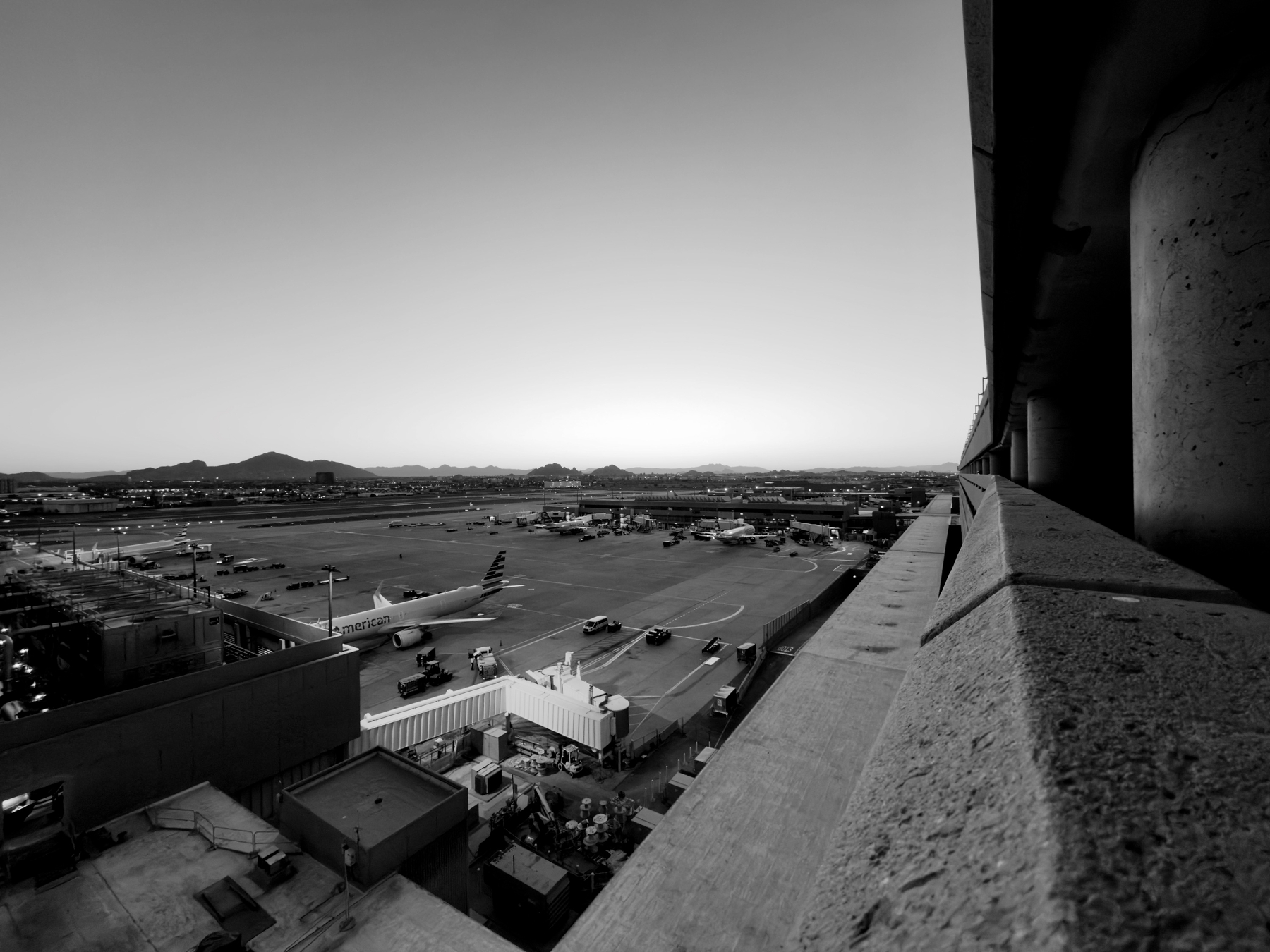 Auto-generated description: A black and white photograph captures an airport tarmac with airplanes, vehicles, and distant hills under an expansive sky.