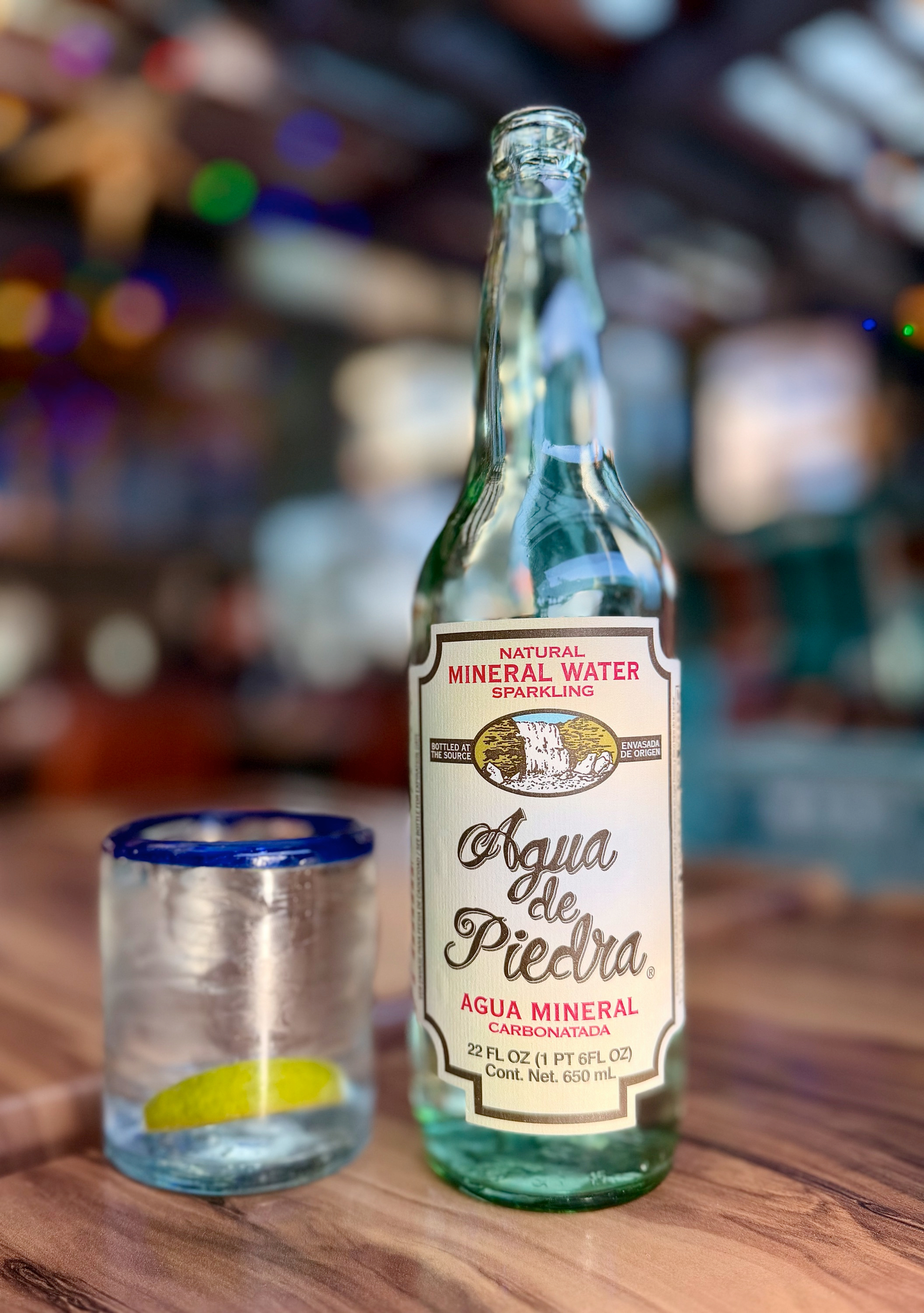 Auto-generated description: A bottle of sparkling mineral water labeled Aqua de Piedra stands on a table next to a glass with a slice of lime.