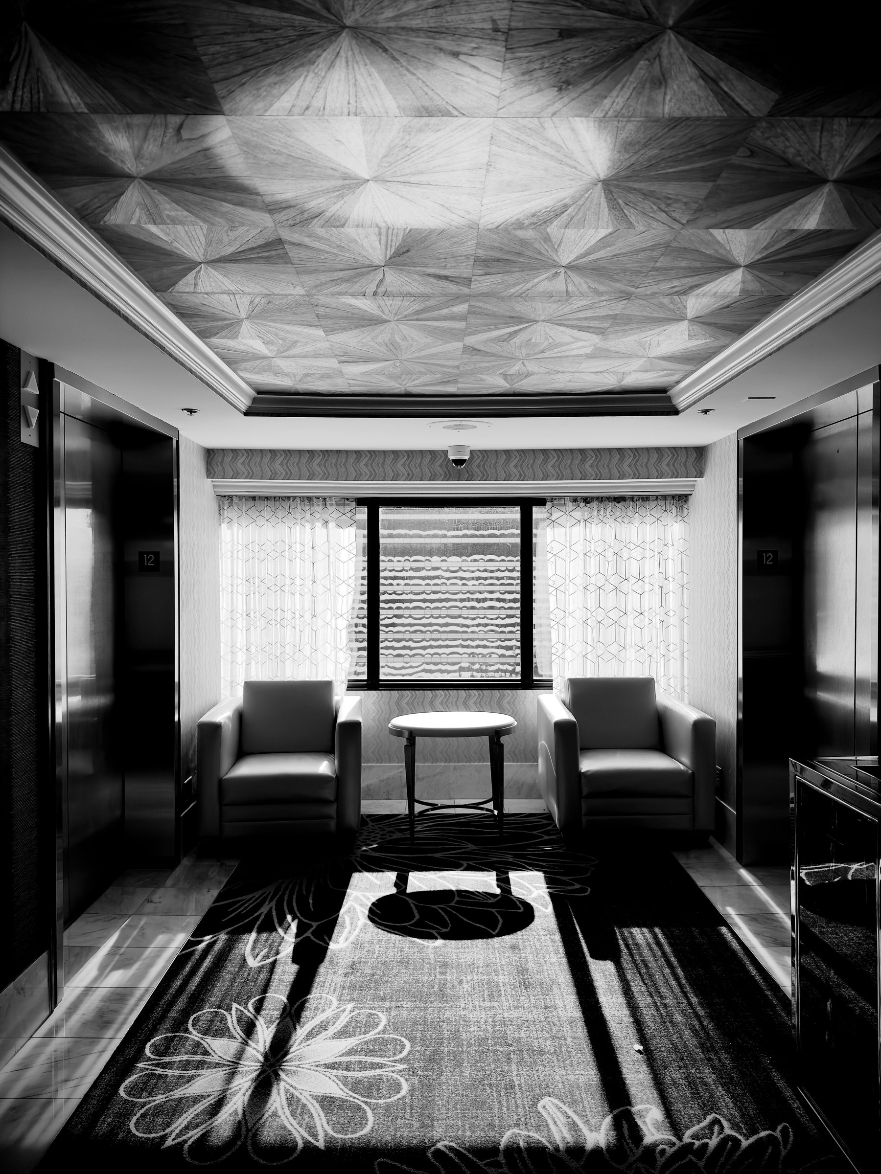Auto-generated description: A symmetric seating area featuring two armchairs and a small round table is positioned between two elevator doors in a hallway illuminated by natural light streaming through a window.