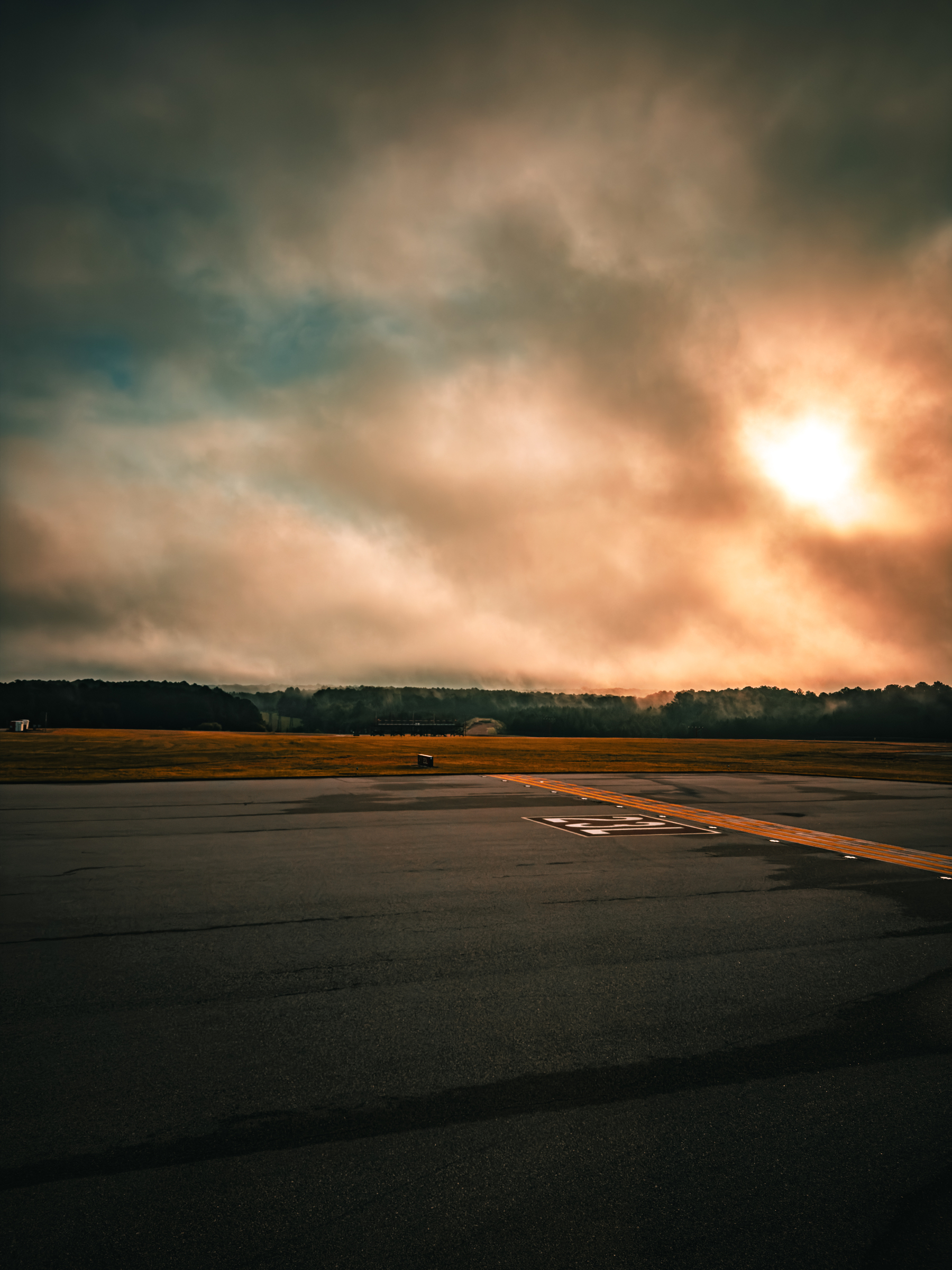 Auto-generated description: A runway or paved road is shown under a cloudy sky with the sun partially visible, casting a warm glow over a distant tree line and grassy area.