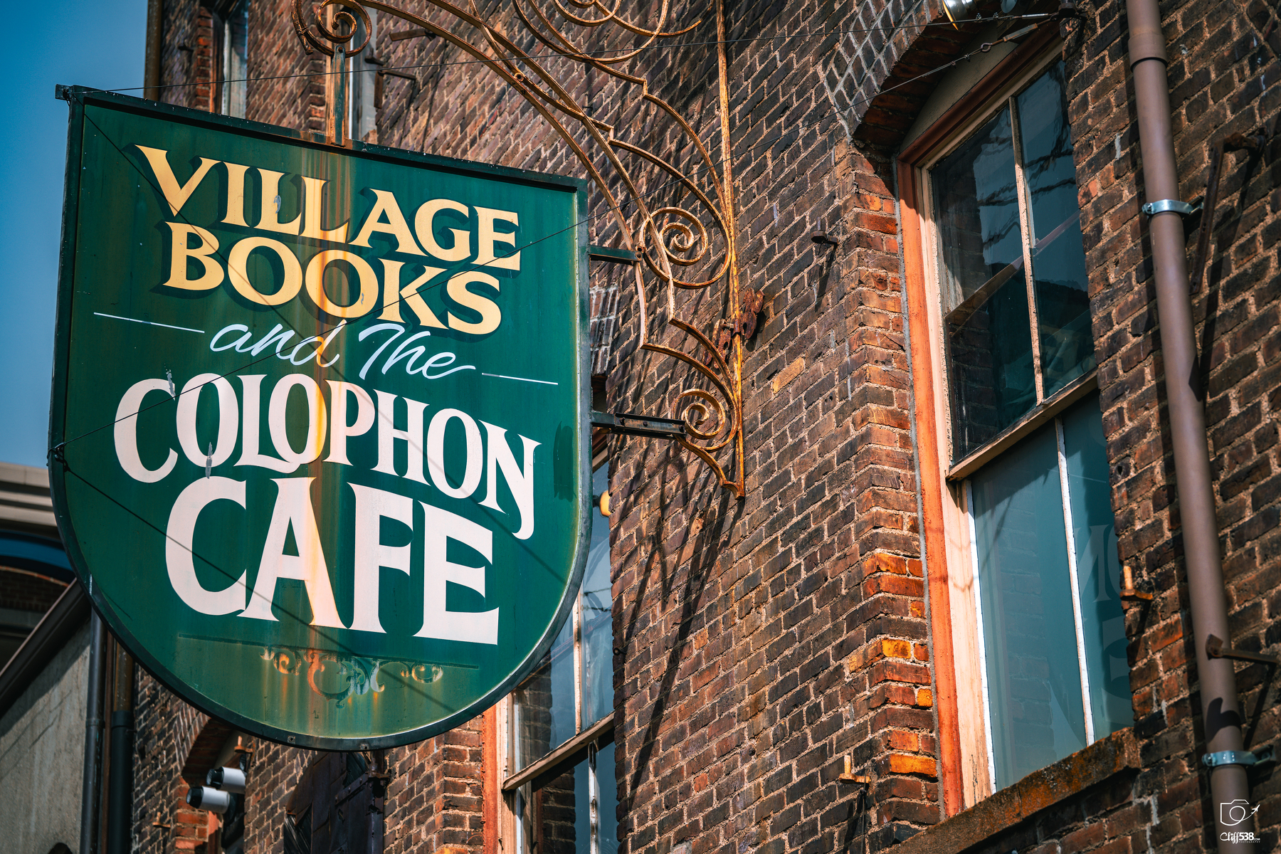 Auto-generated description: A green sign with yellow and white text hangs outside a brick building, advertising Village Books and The Colophon Cafe.