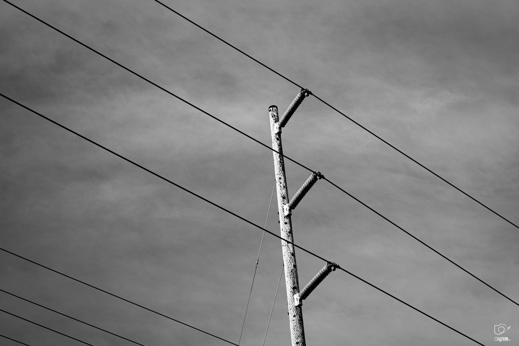 Auto-generated description: A wooden utility pole with multiple power lines stretches across a cloudy sky in a black and white photograph.