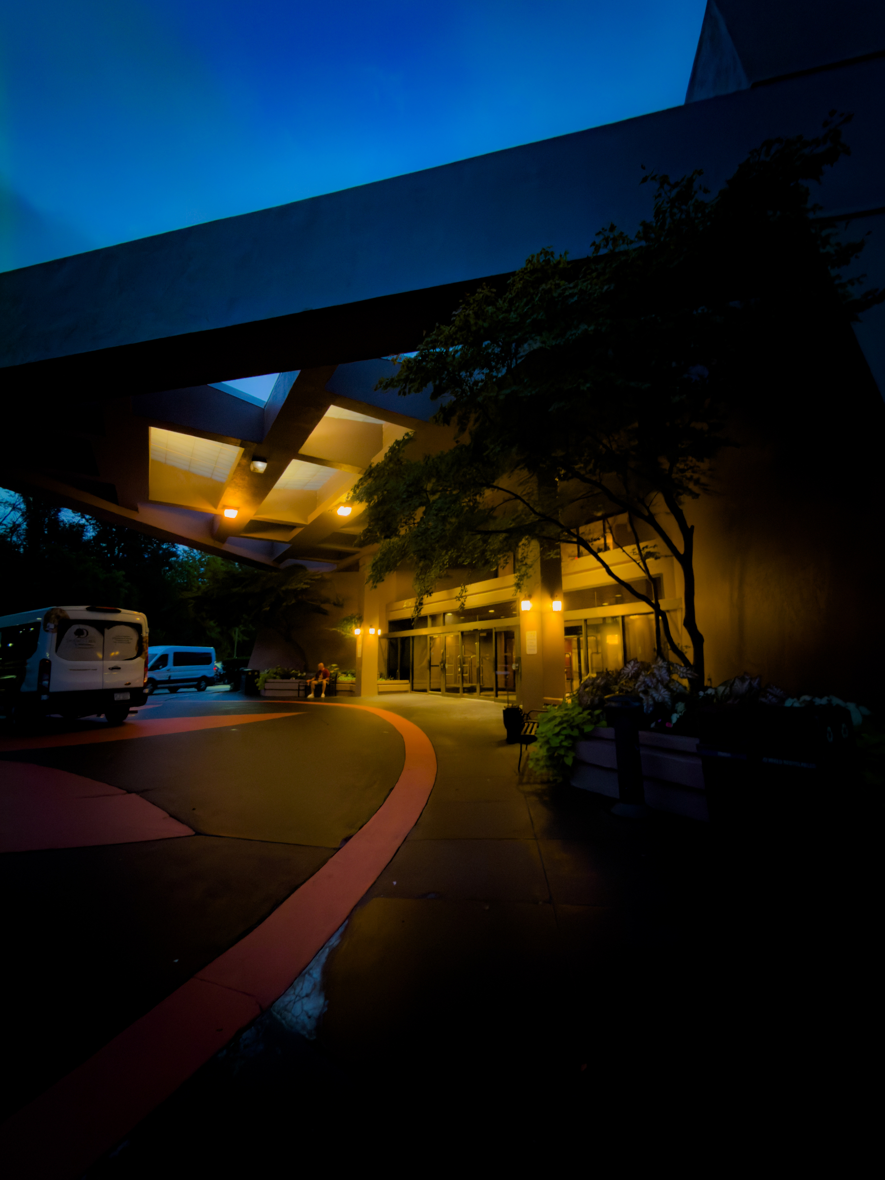 Auto-generated description: A dimly lit building entrance with overhanging structures, illuminated by yellow lights, is surrounded by trees and parked vans during twilight.