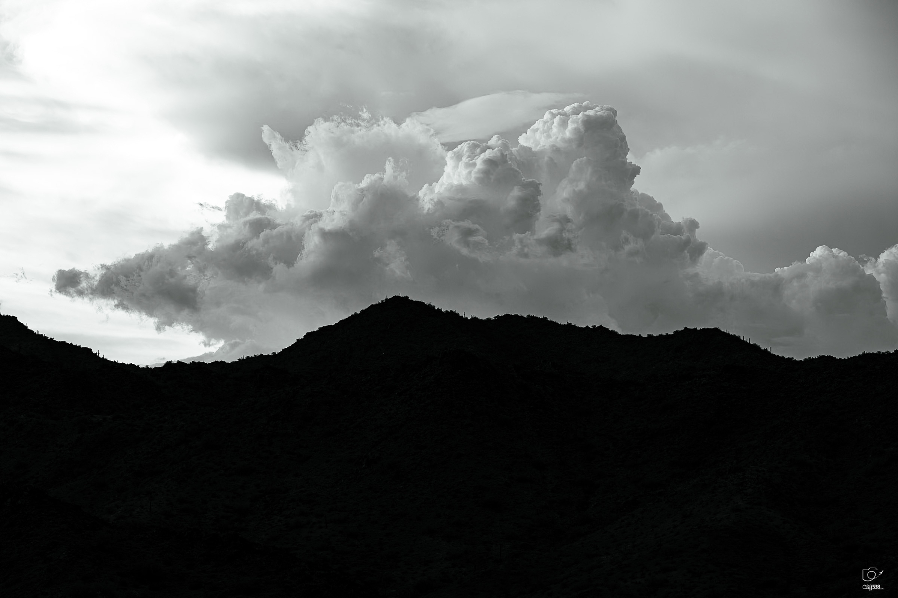 Auto-generated description: A large, billowing cloud formation looms above the silhouette of a mountain range under a cloudy sky.