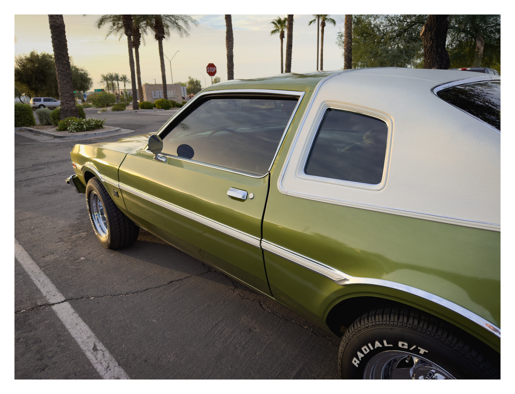 Auto-generated description: A green vintage car with white detailing is parked in a lot near palm trees at sunset.