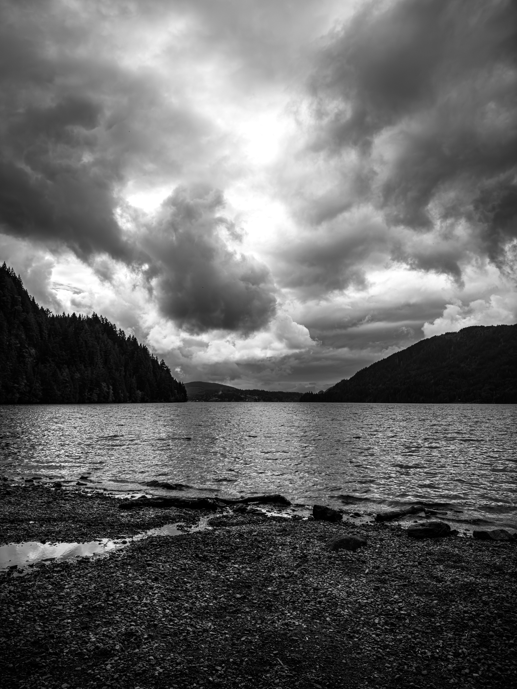 Auto-generated description: A serene lake is surrounded by tree-covered hills under a dramatic, cloudy sky.