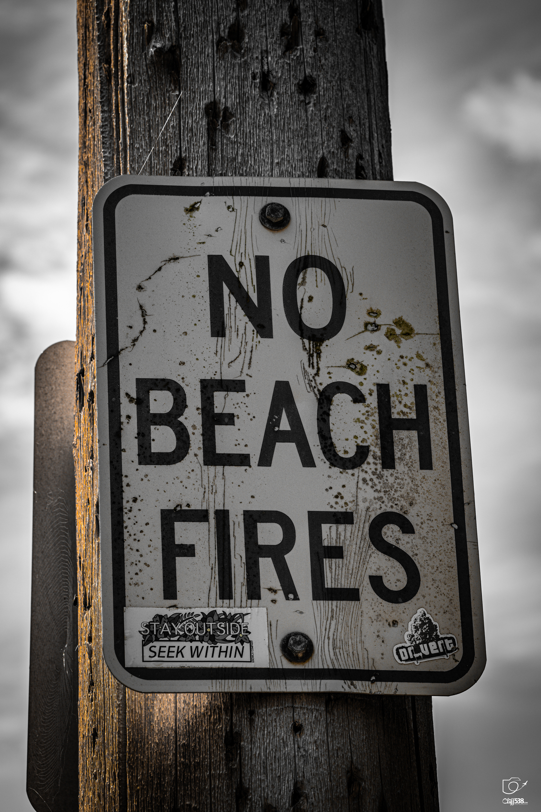 Auto-generated description: A weathered sign attached to a wooden post forbids beach fires and is marked with stickers, set against a cloudy sky.